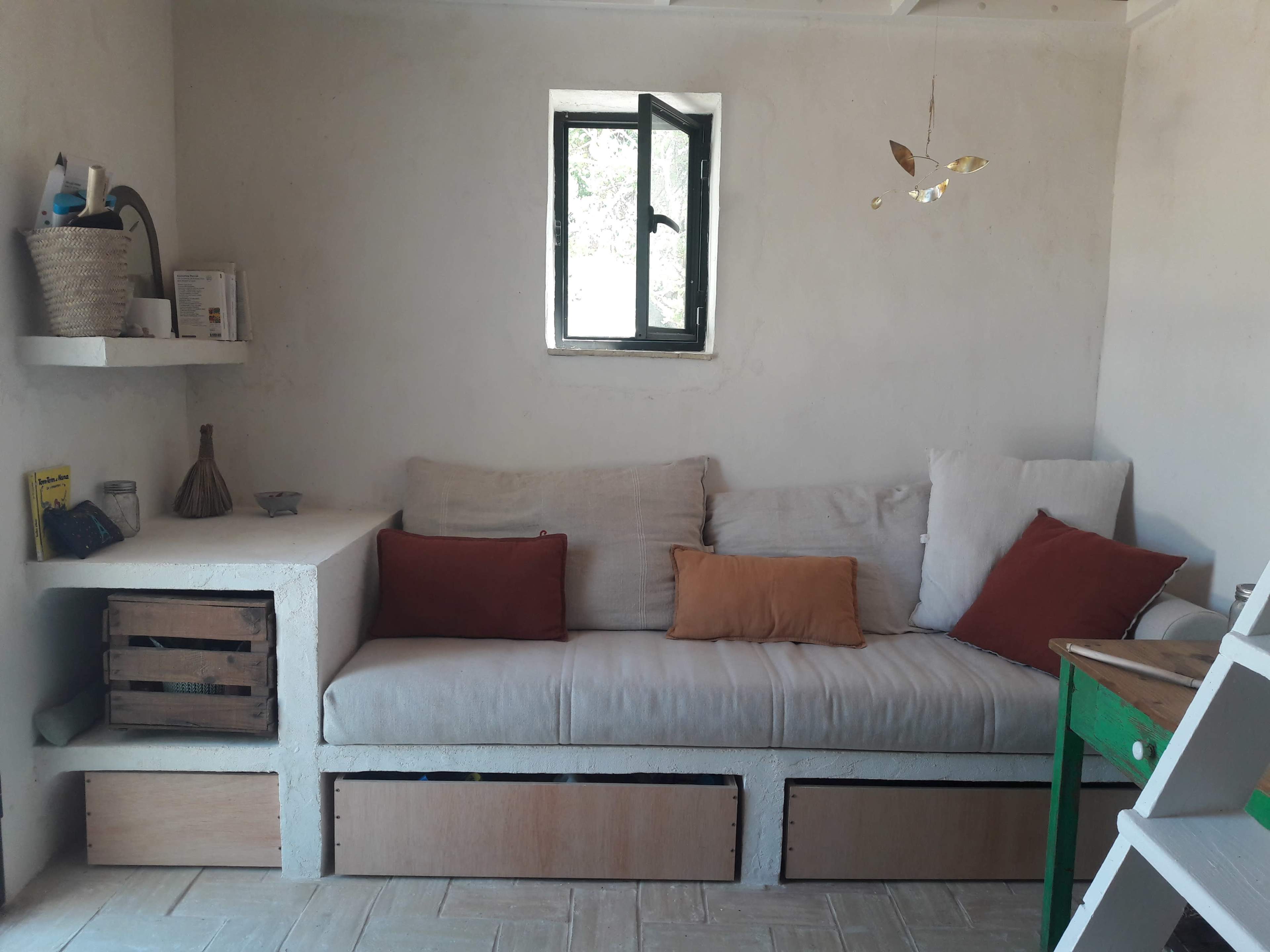 A minimalist living space features a light-colored sofa with decorative throw pillows, a small shelf with items, and a window that provides natural light.