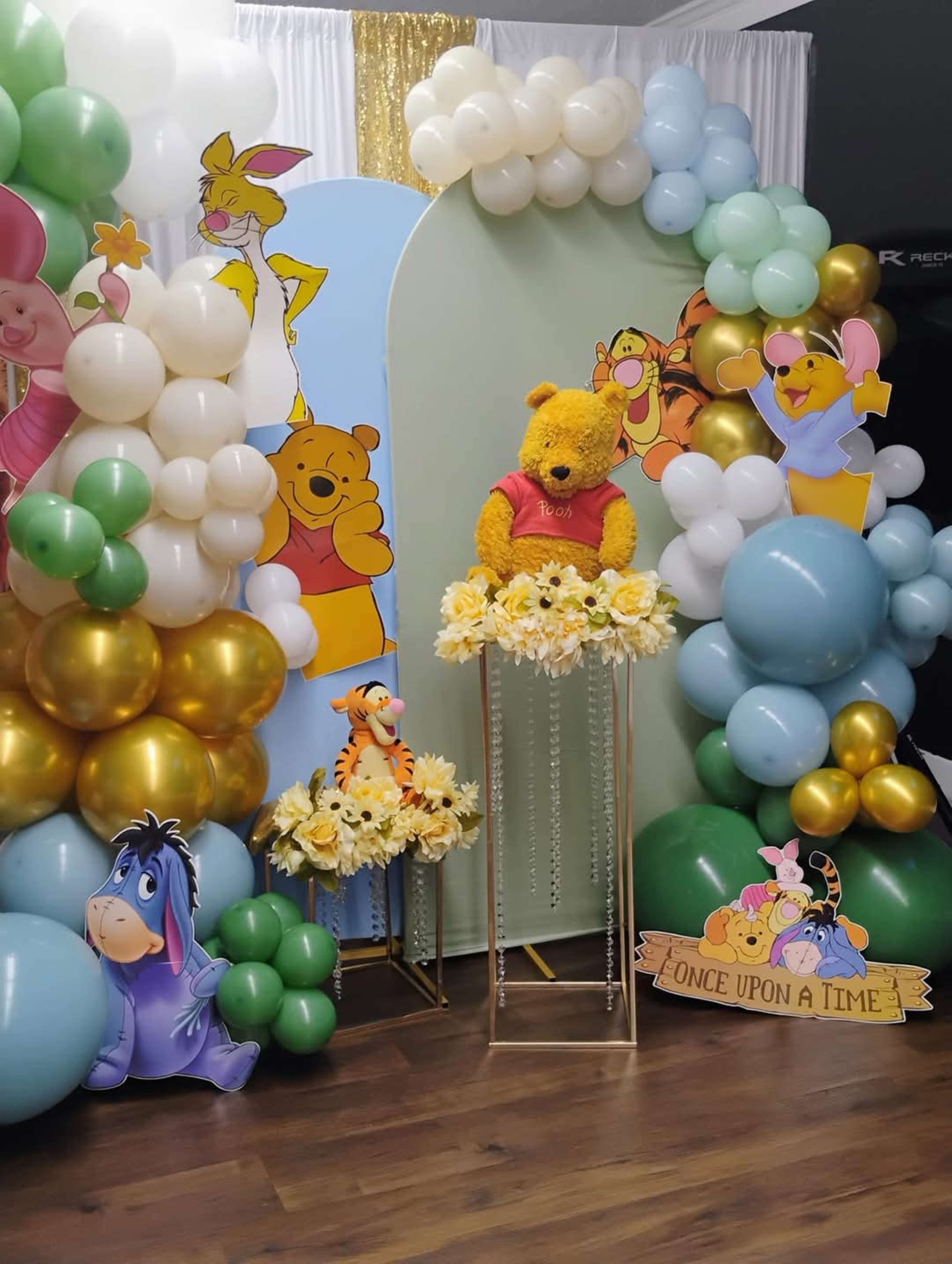 The image features a colorful balloon arch setup with a plush teddy bear dressed as Winnie the Pooh and cartoon character cutouts, including Tigger, Eeyore, and Piglet, arranged around a decorative display.