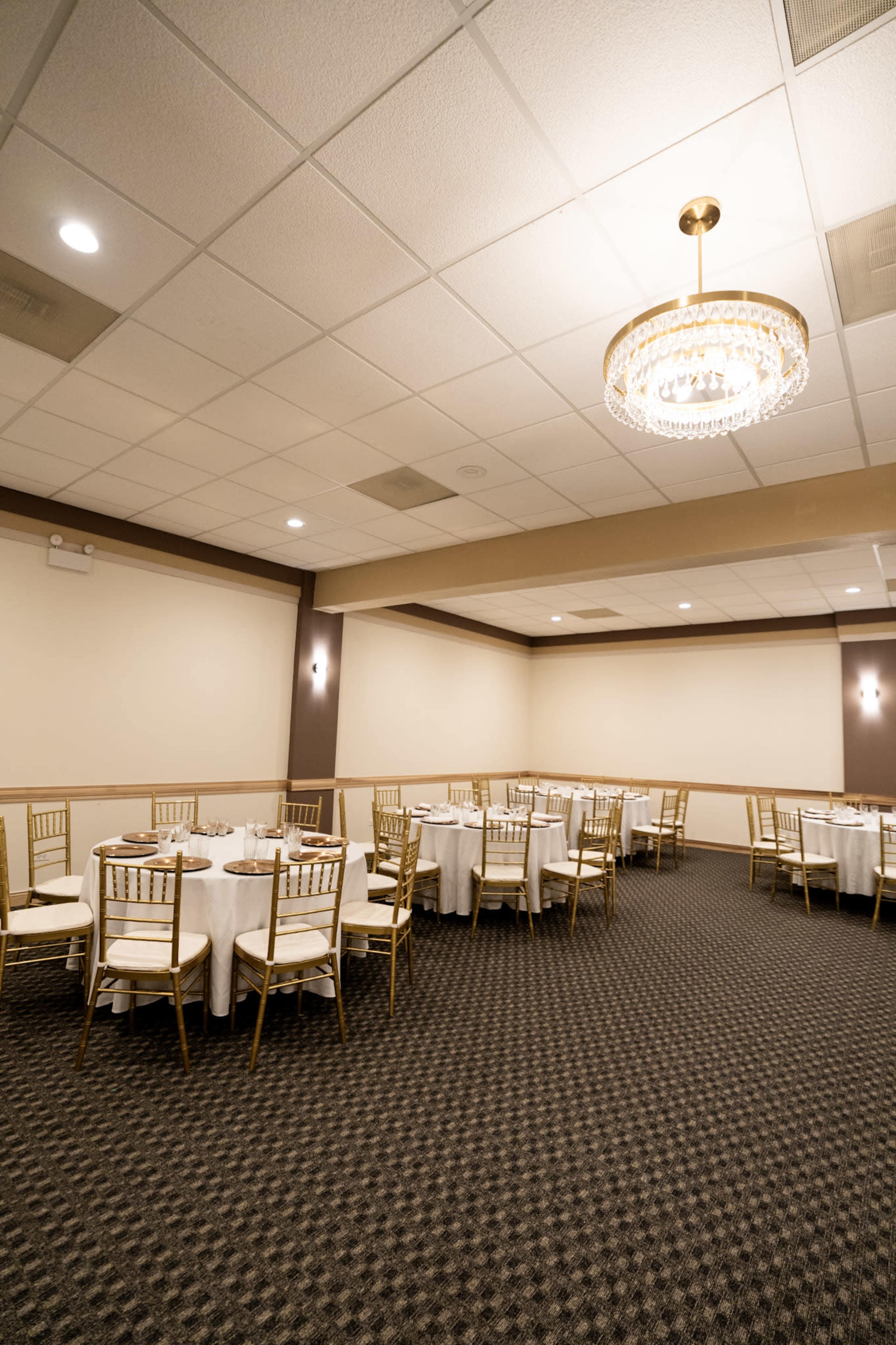 The image shows a spacious, well-lit banquet room with round tables covered in white tablecloths and gold chairs arranged neatly on a patterned carpet.