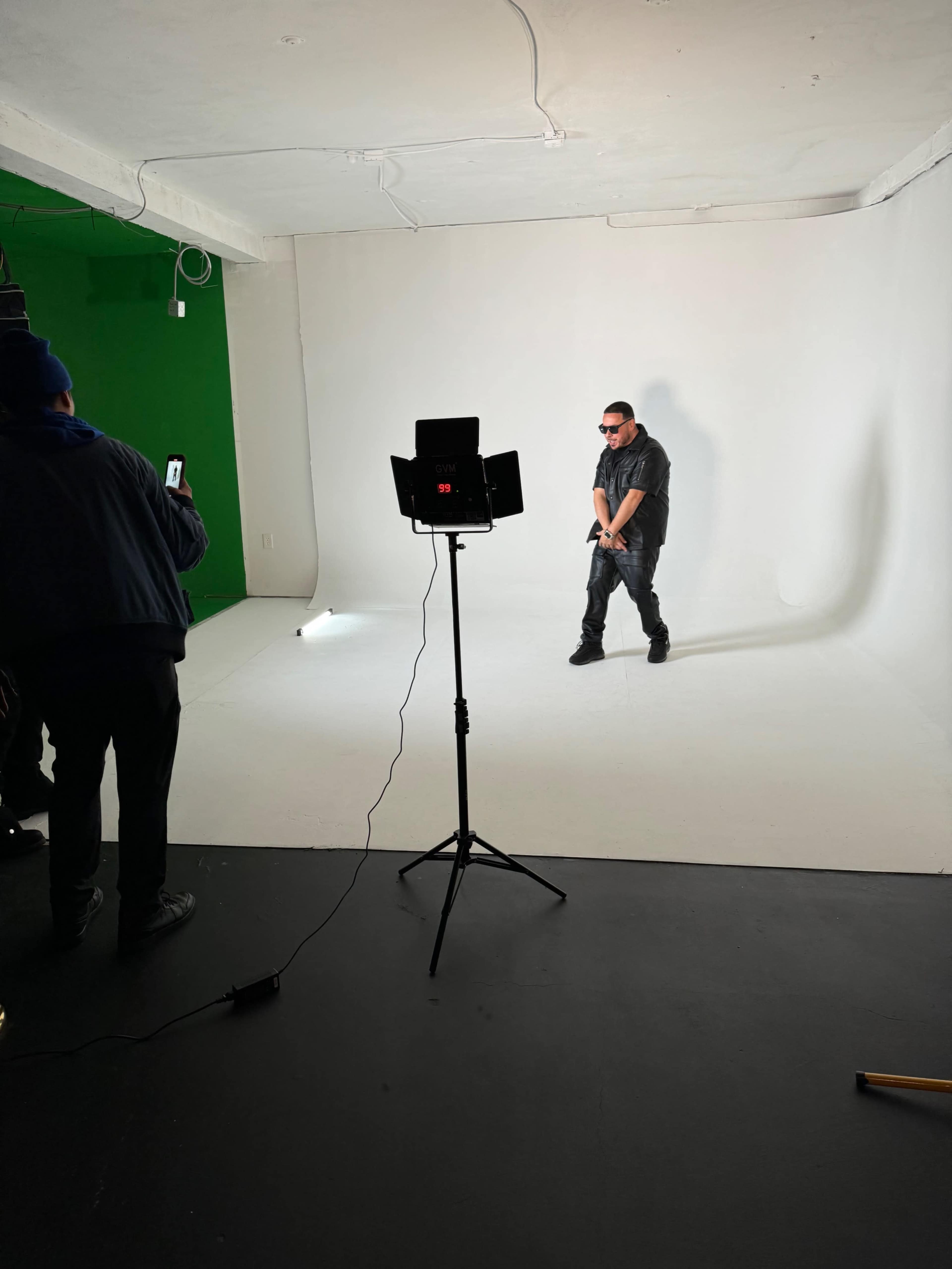 A person stands in a white studio space while another person takes photos from the side, with a green wall visible in the background.
