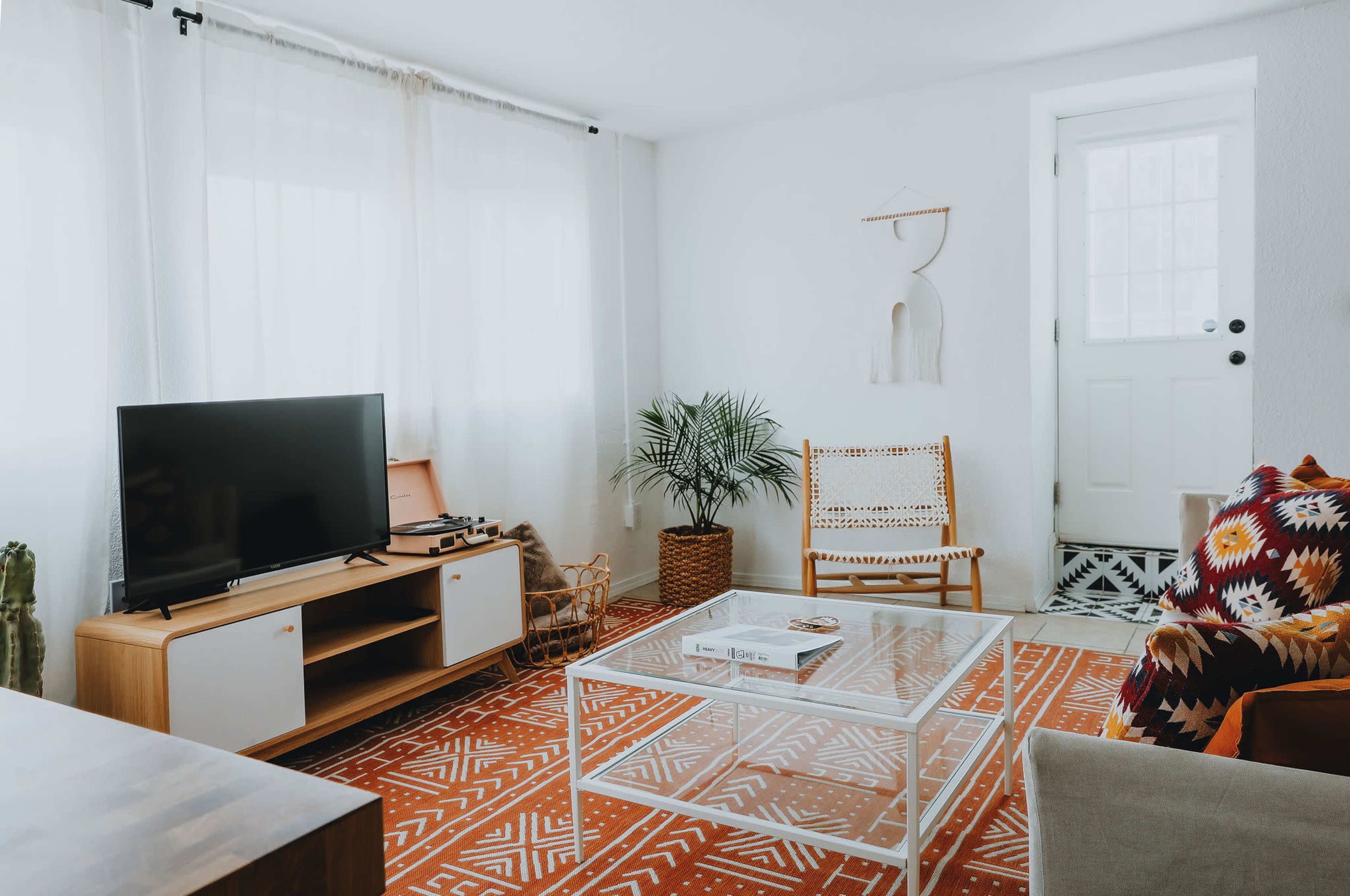 A modern living room features a television on a wooden media console, a glass coffee table on a patterned rug, and a chair beside a door with natural light streaming in through sheer curtains.