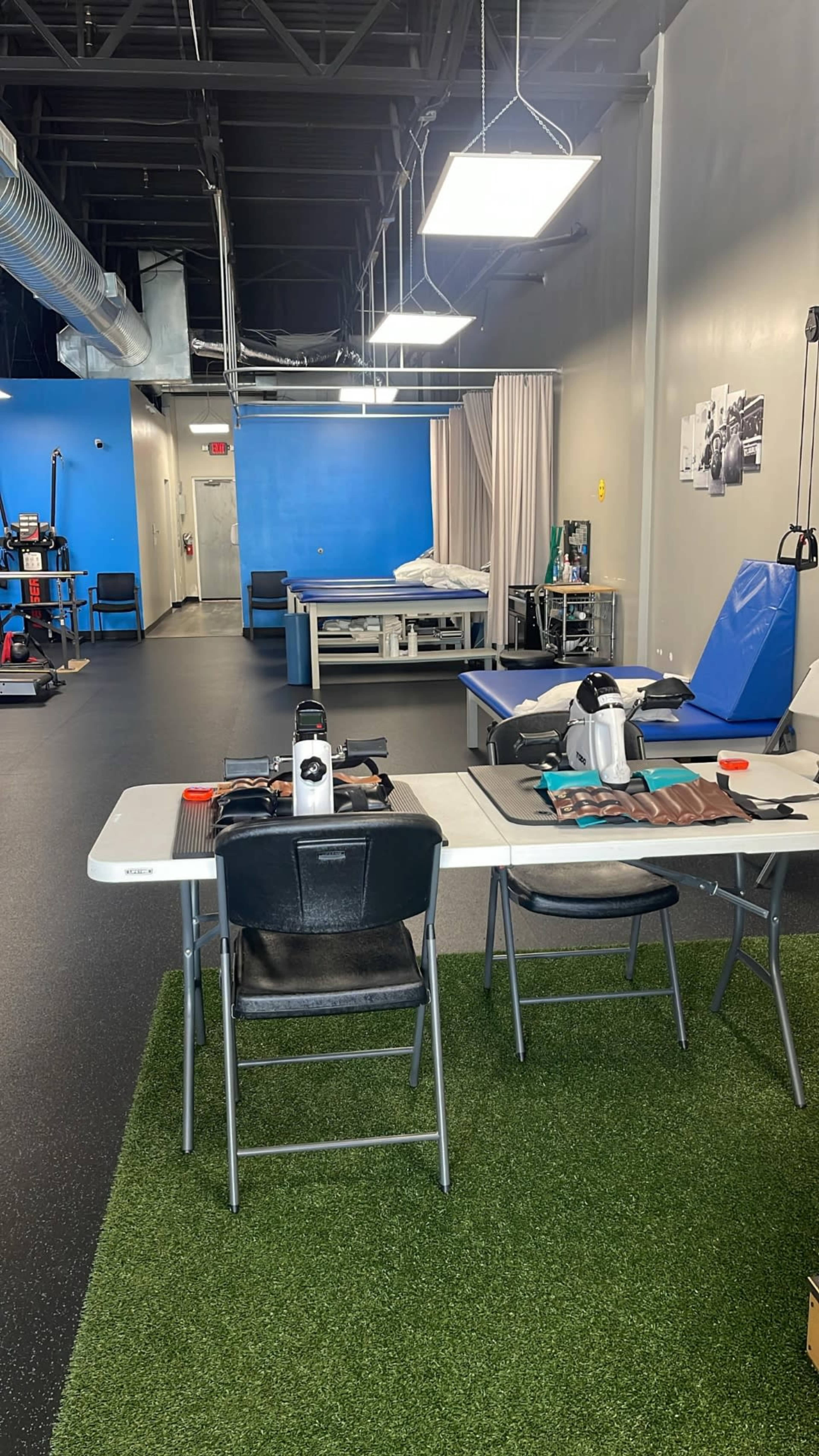 The image shows a spacious therapy room with blue walls, tables for treatment, and equipment arranged on a carpet-like surface.