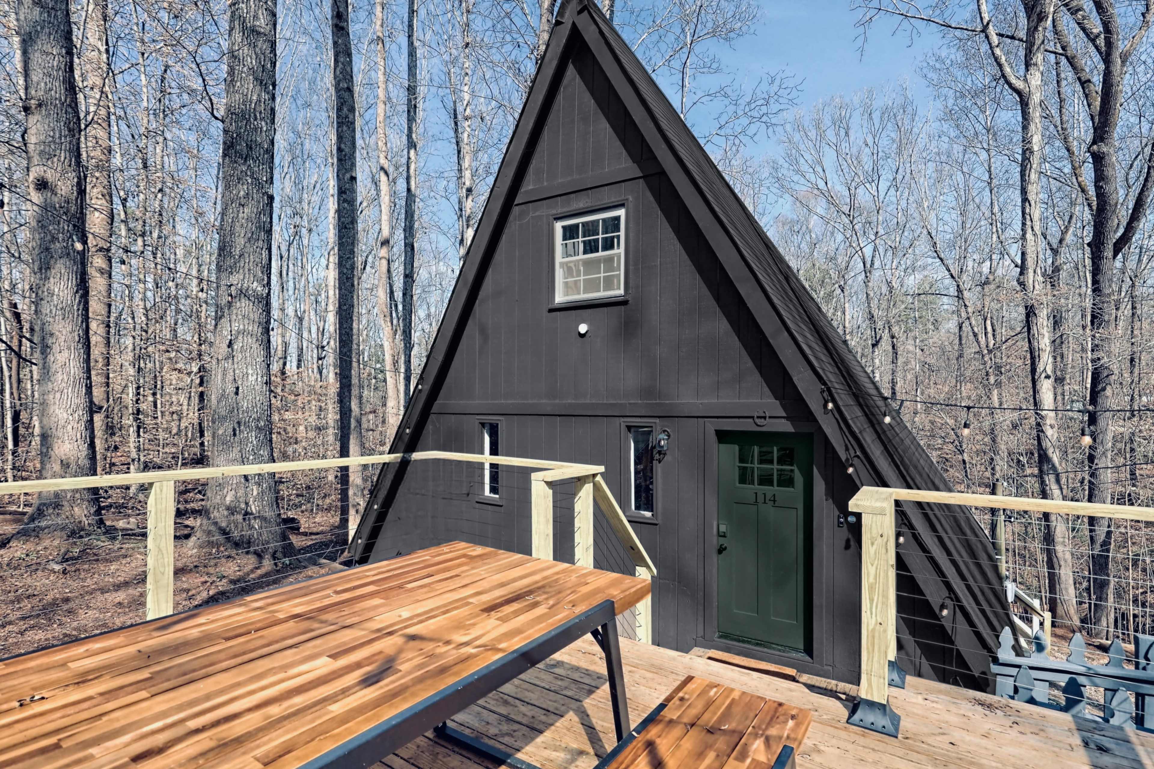 A dark A-frame cabin stands in a wooded area, with a wooden deck and picnic table in the foreground.
