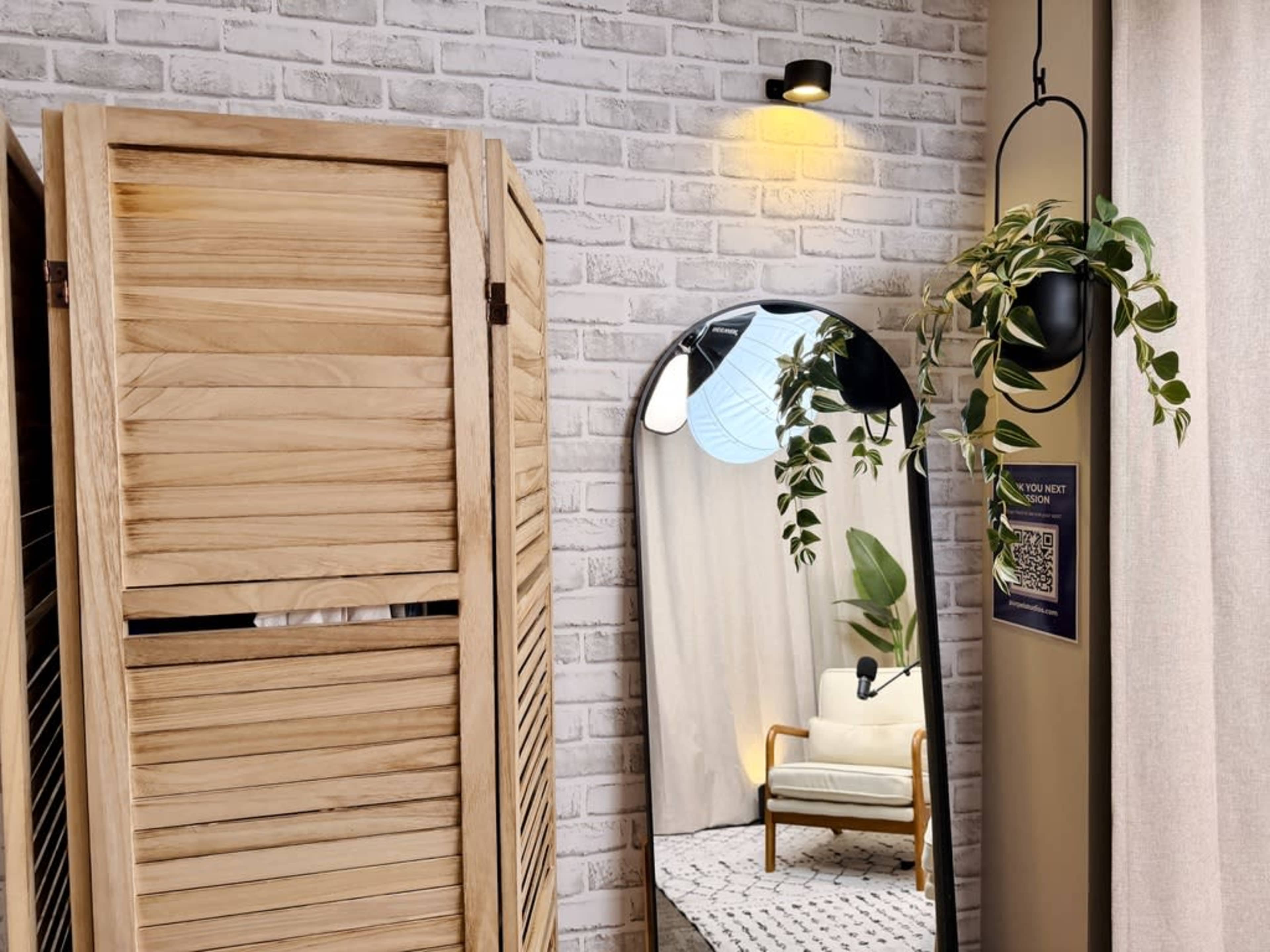 The image shows a corner of a modern interior space featuring a wooden privacy screen, a large mirror, a hanging plant, and a chair in front of light-colored curtains and a textured wall.