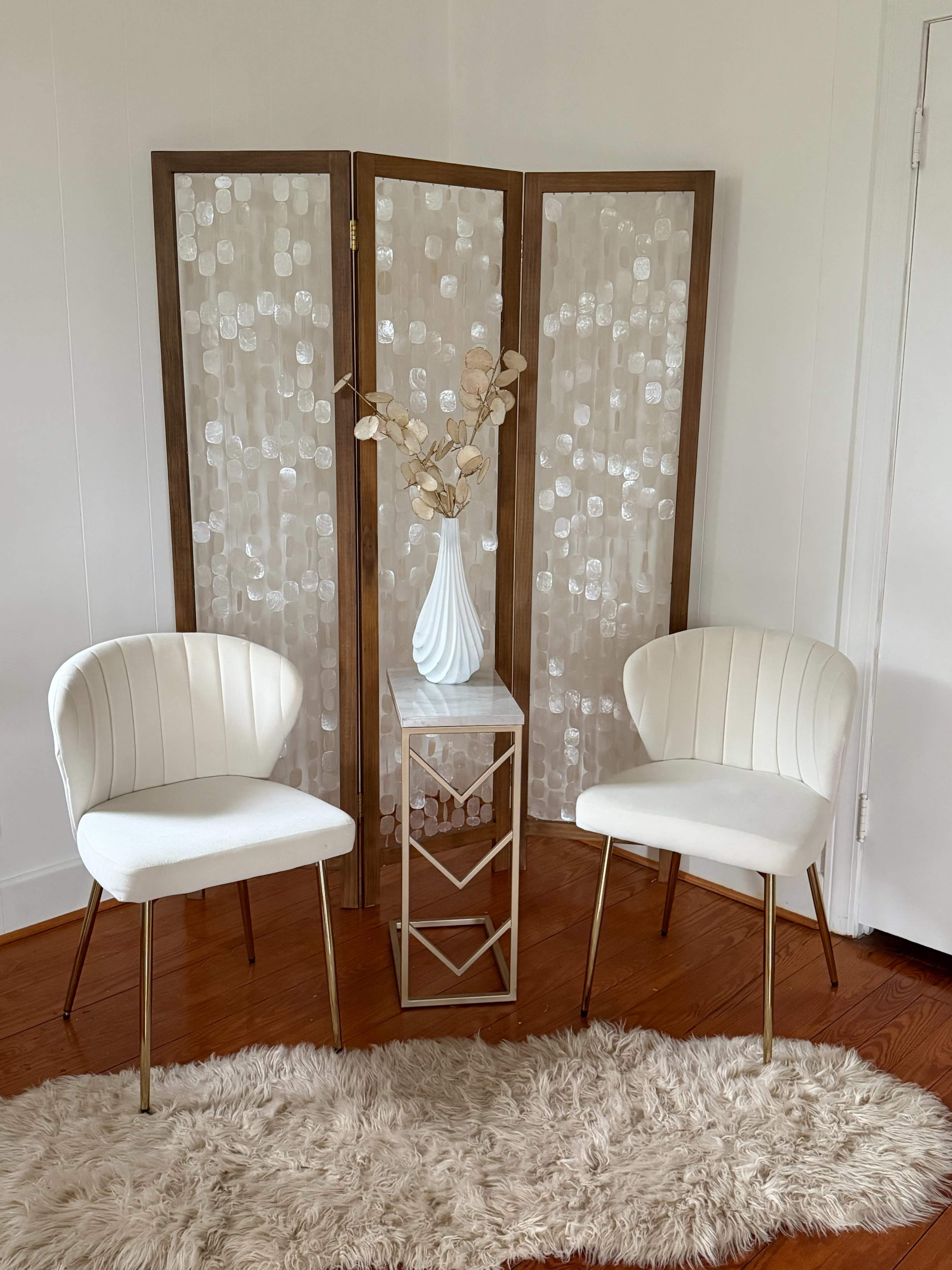 The image shows a cozy interior scene featuring two white chairs, a small glass side table, a decorative vase with dried branches, and a folding screen in the background, all set on a fluffy rug.