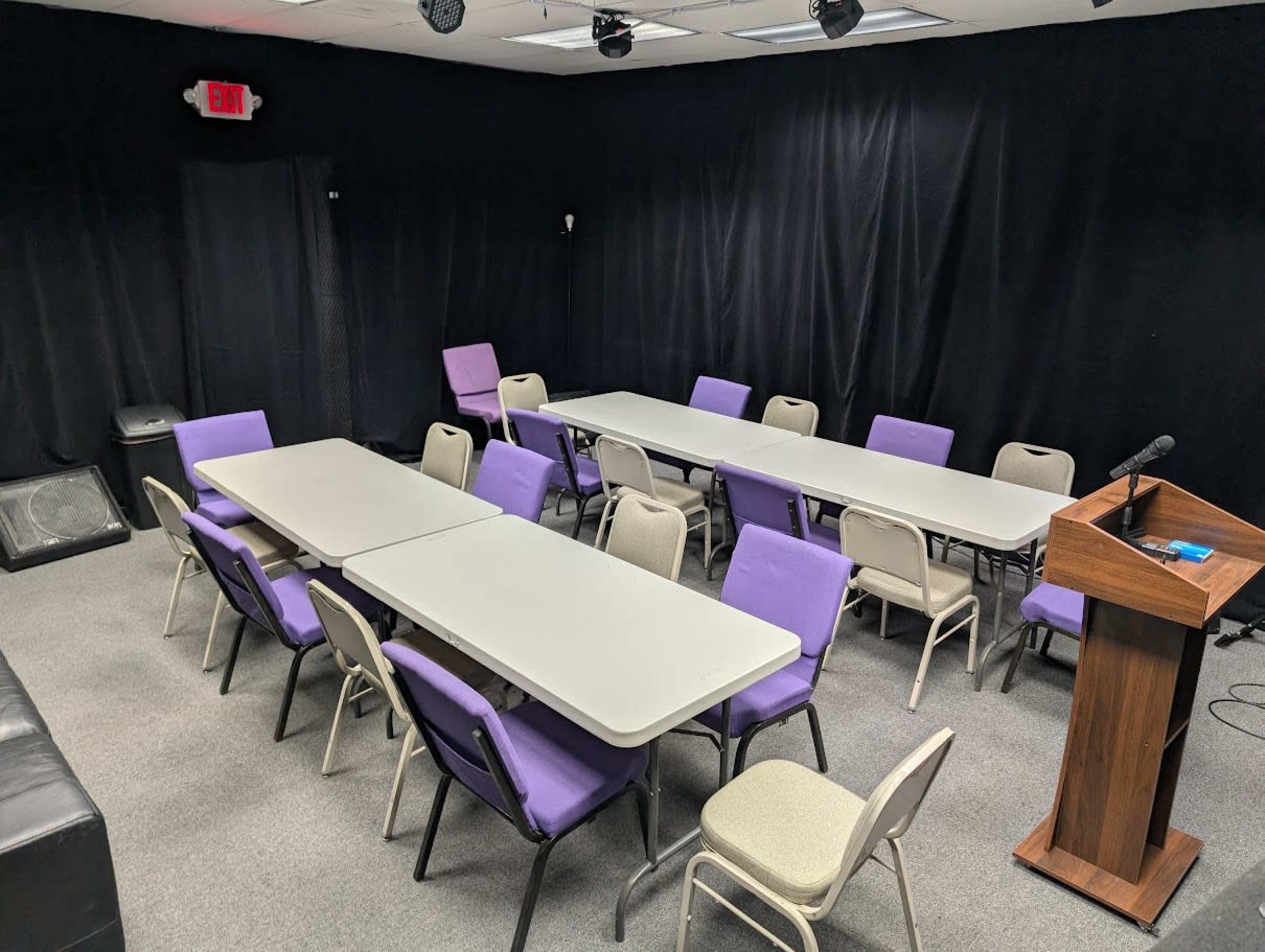 The image shows a conference room with multiple tables arranged in a U-shape, surrounded by purple and beige chairs, and a podium at one end.