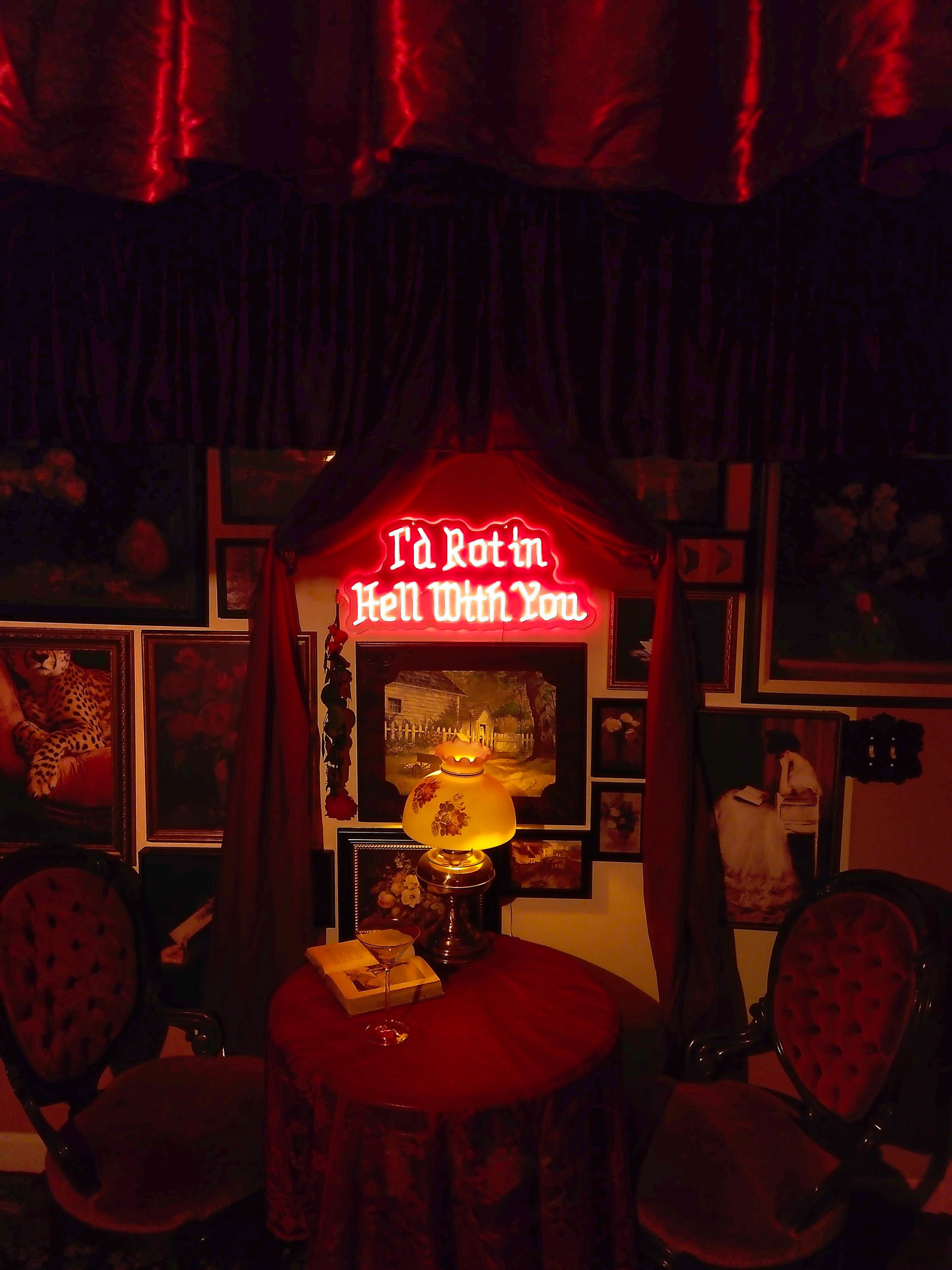The image depicts a dimly lit room adorned with various framed artworks, featuring a neon sign that reads "I'd Rot in Hell With You" over a small table with a lamp and two chairs.