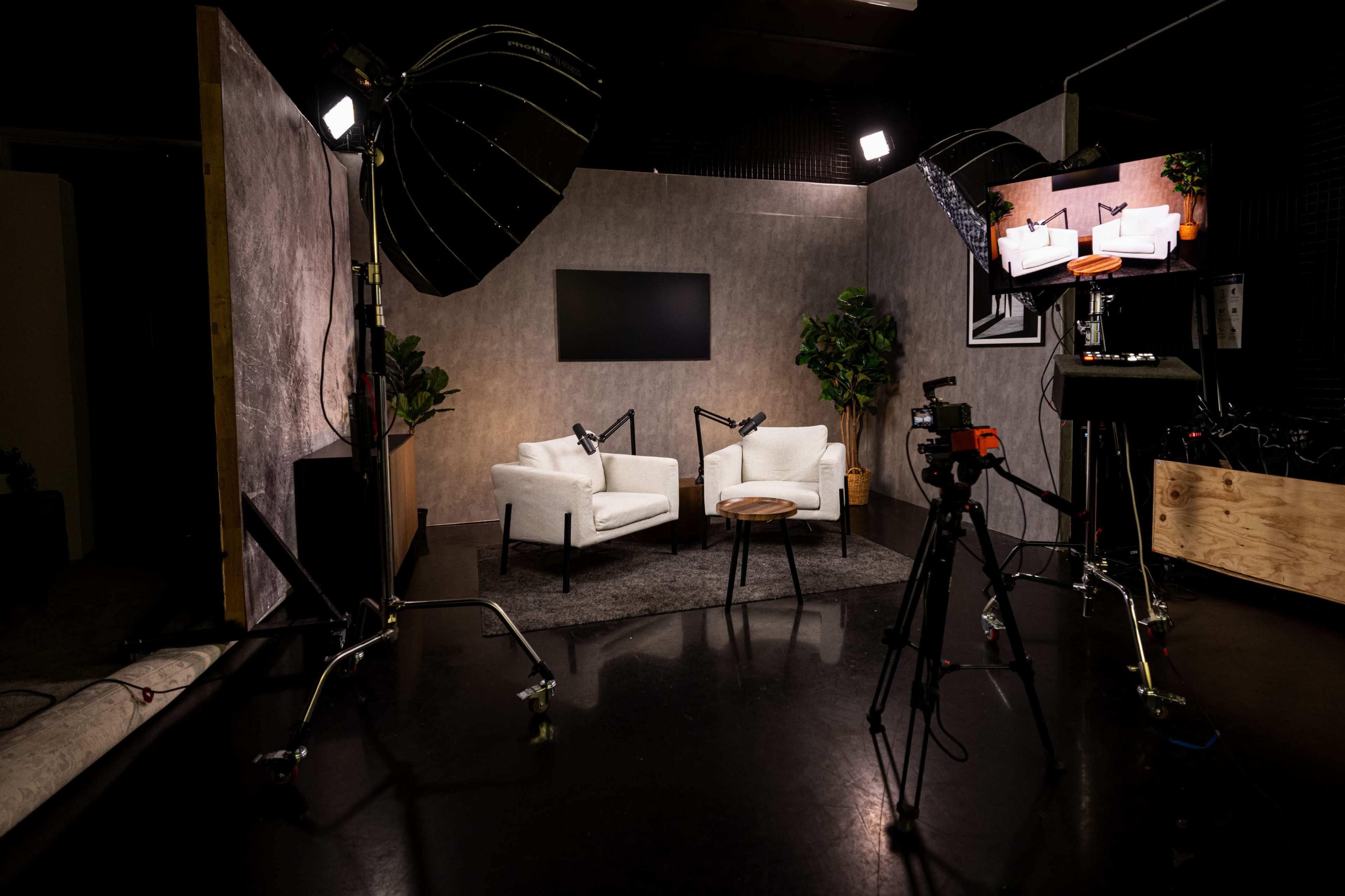 The image shows a professional studio setup featuring two white armchairs, a wooden side table, a backdrop, and lighting equipment ready for filming.