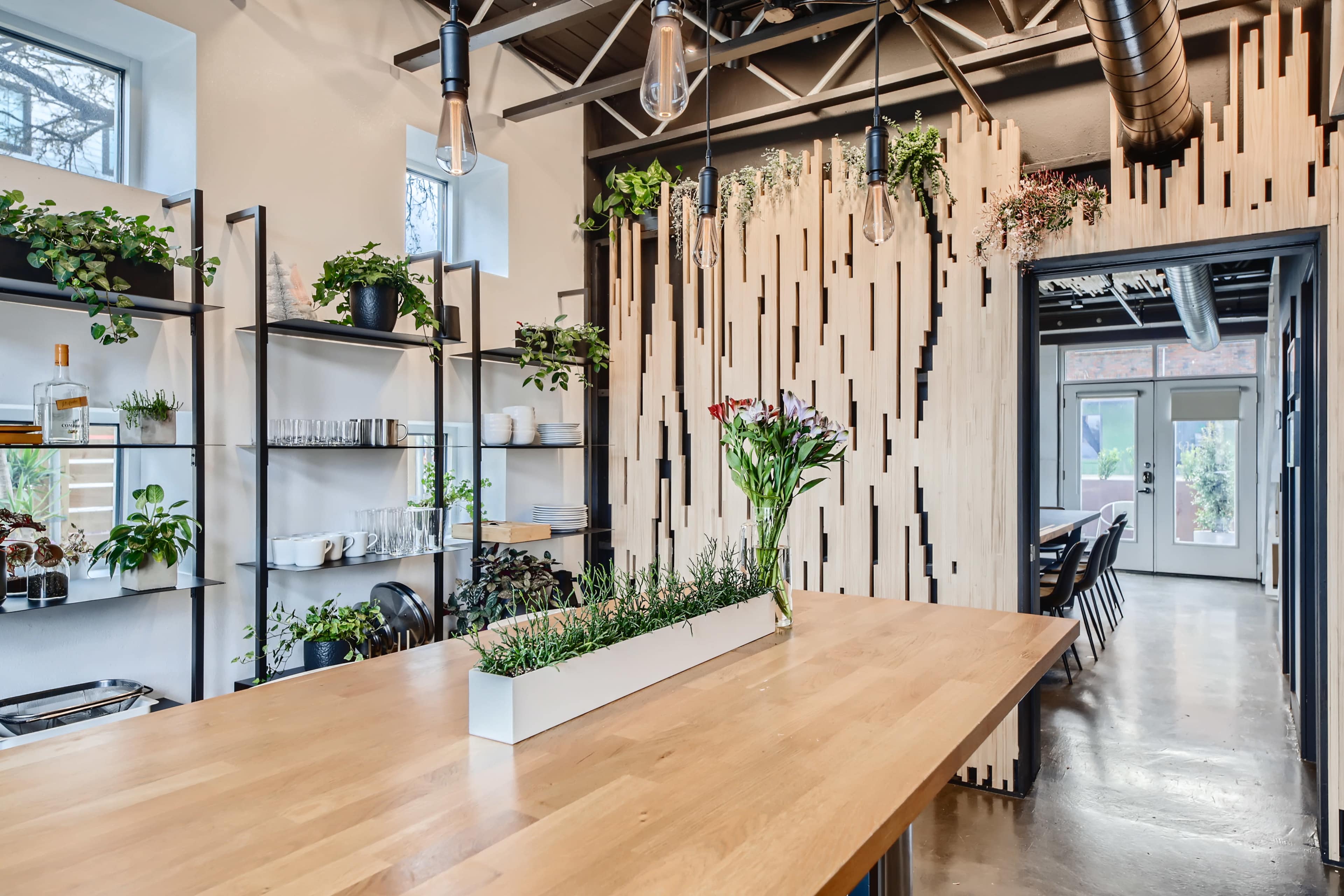 A modern kitchen space featuring wooden shelves with plants, a large wooden table, and hanging light fixtures.