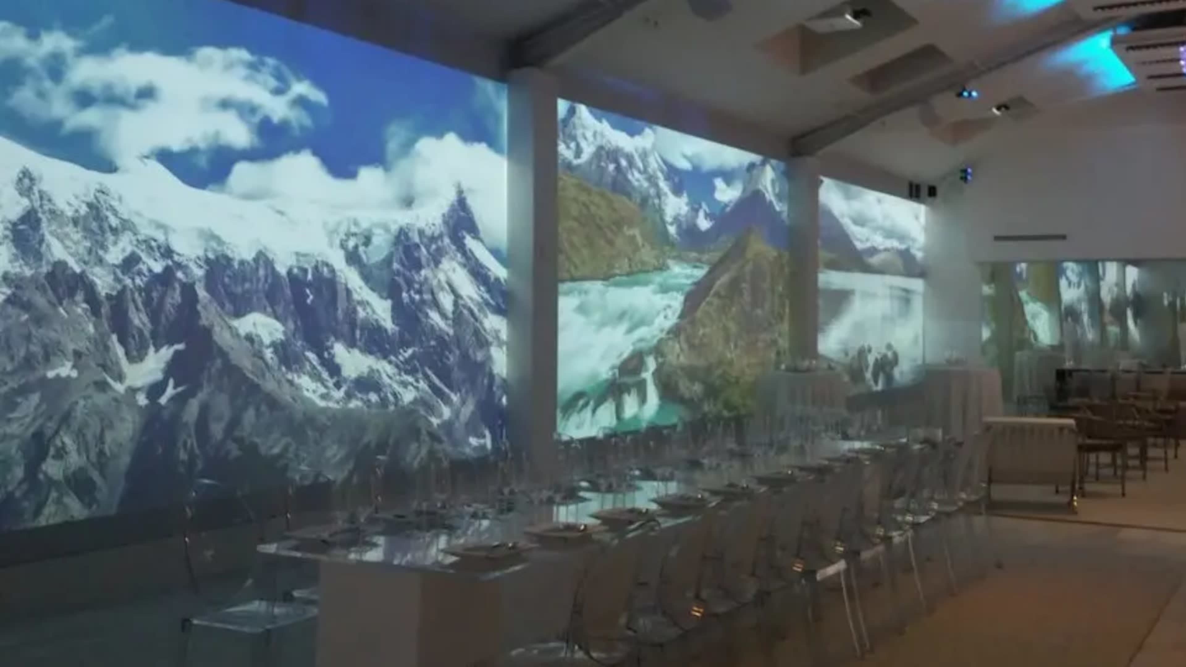 The image shows a modern dining setup with a long table surrounded by transparent chairs, while large-scale projections of mountainous landscapes adorn the walls.