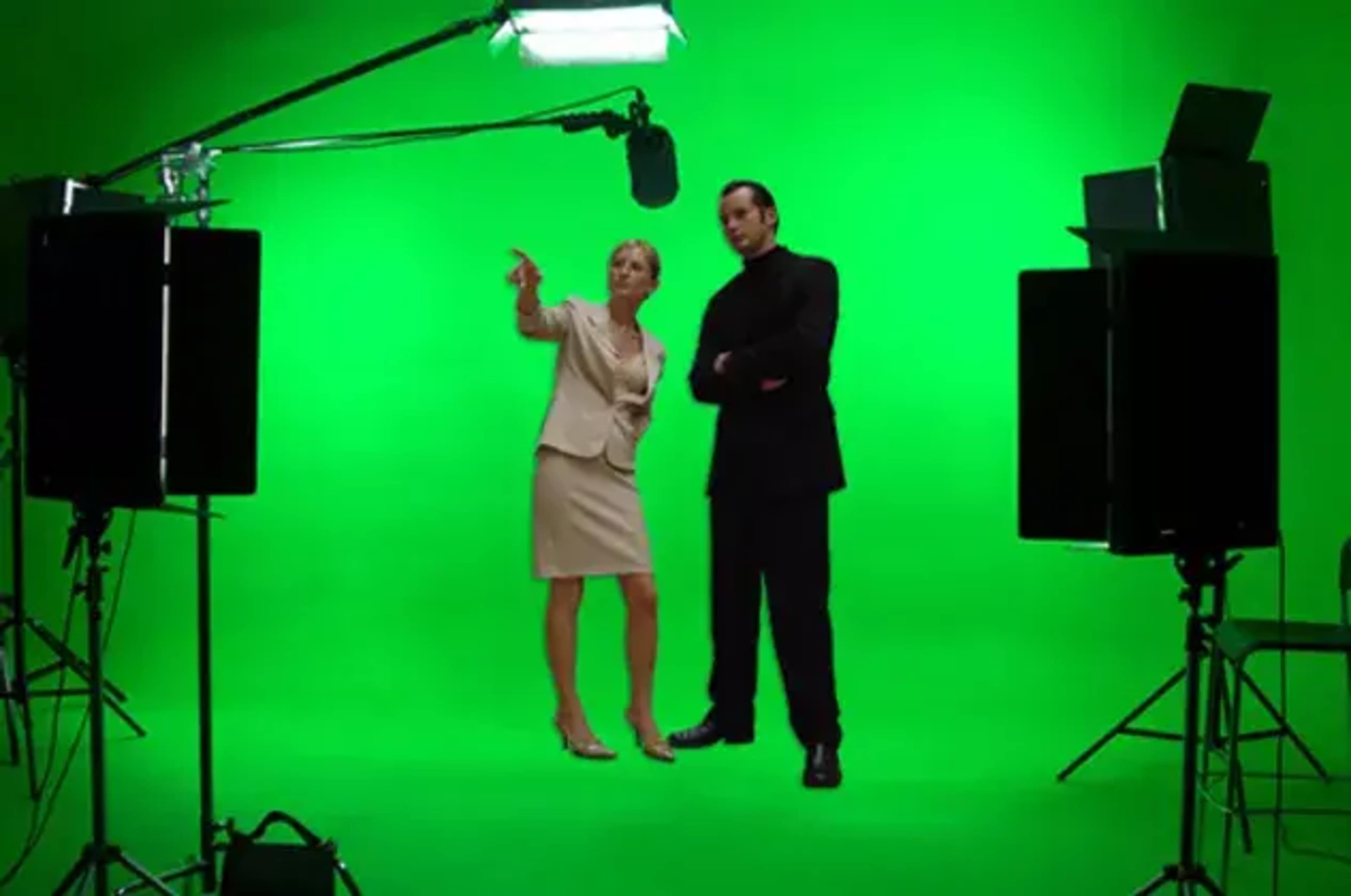 A woman in business attire and a man in black clothing stand on a green screen set, surrounded by studio lighting equipment.