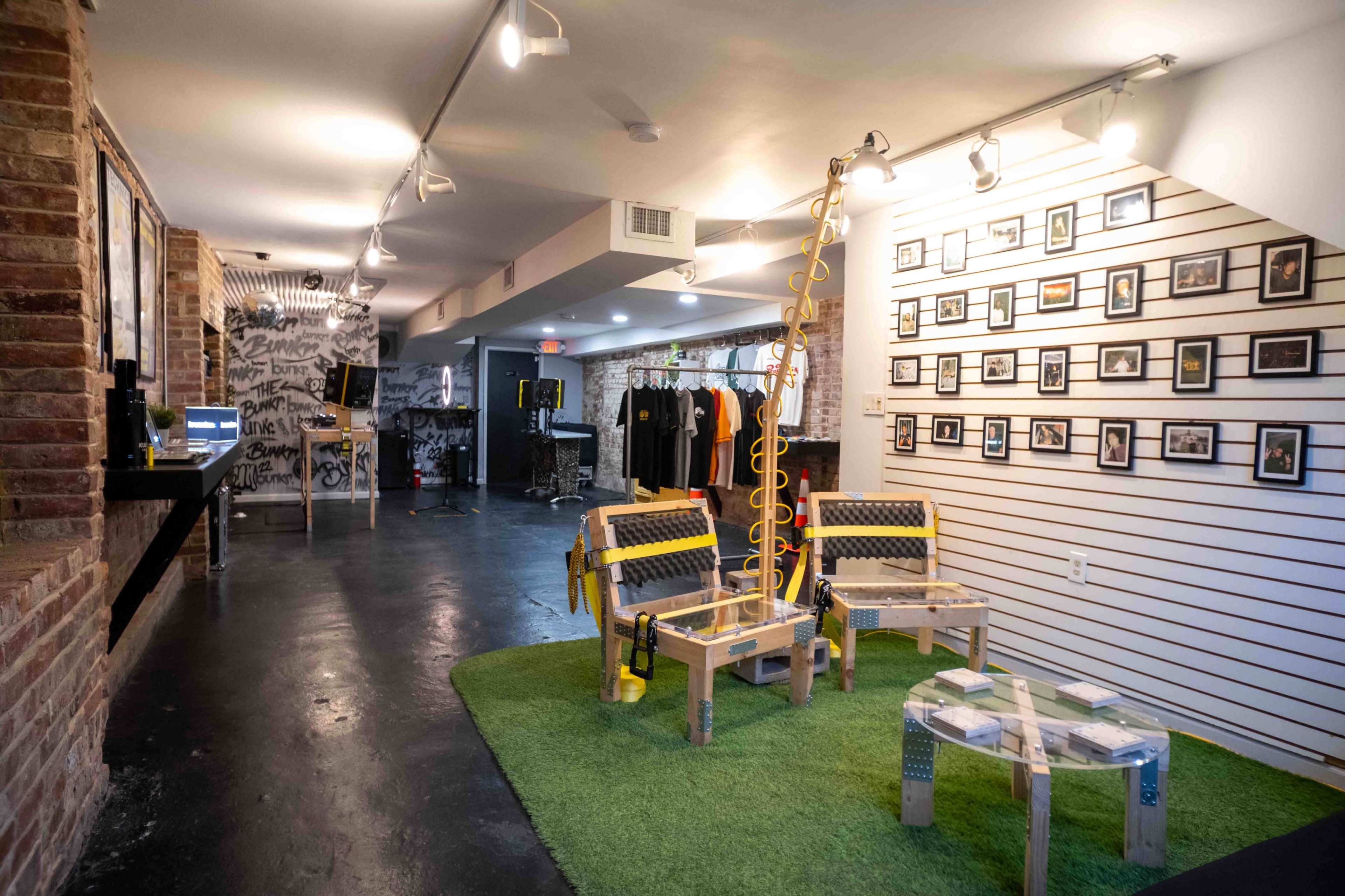 A modern indoor space featuring a seating area with wooden chairs on a green carpet, surrounded by framed photographs on the walls and a display of clothing in the background.