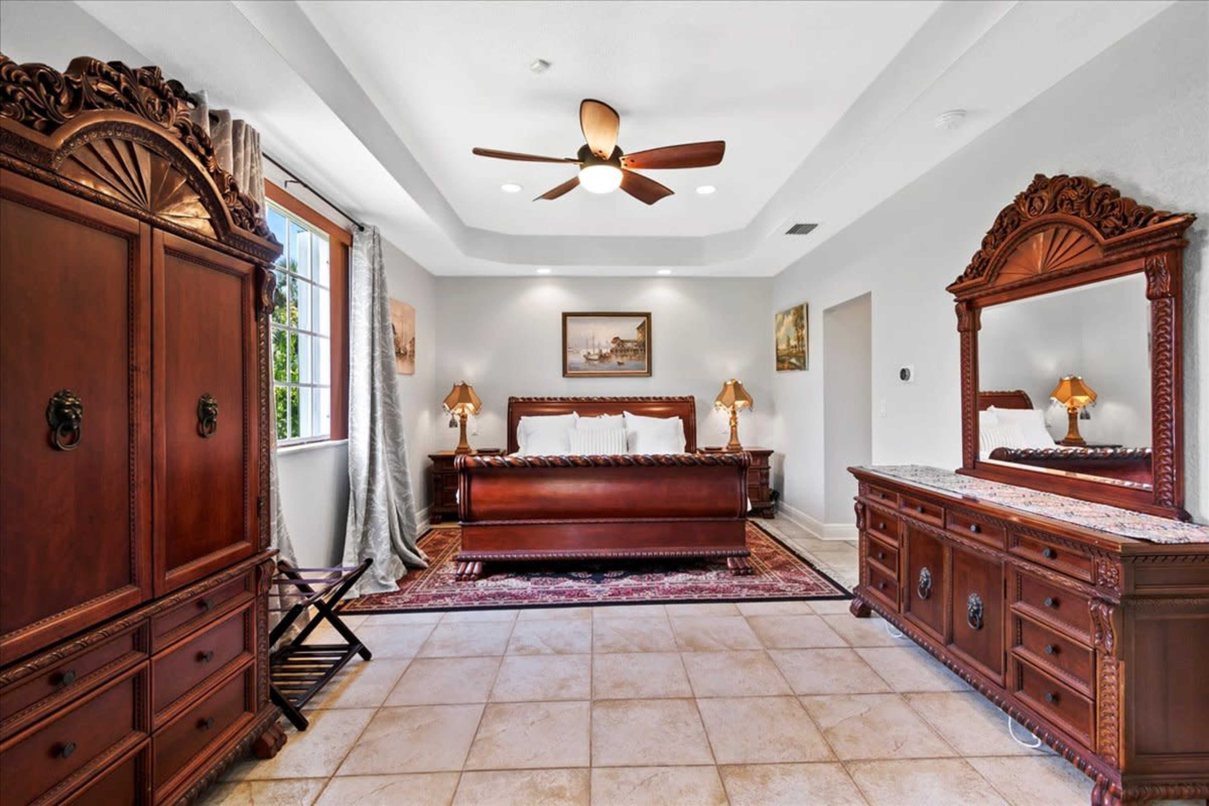 A spacious bedroom featuring ornate wooden furniture, including a bed, dresser, and wardrobe, with beige tile flooring and soft natural light from a window.