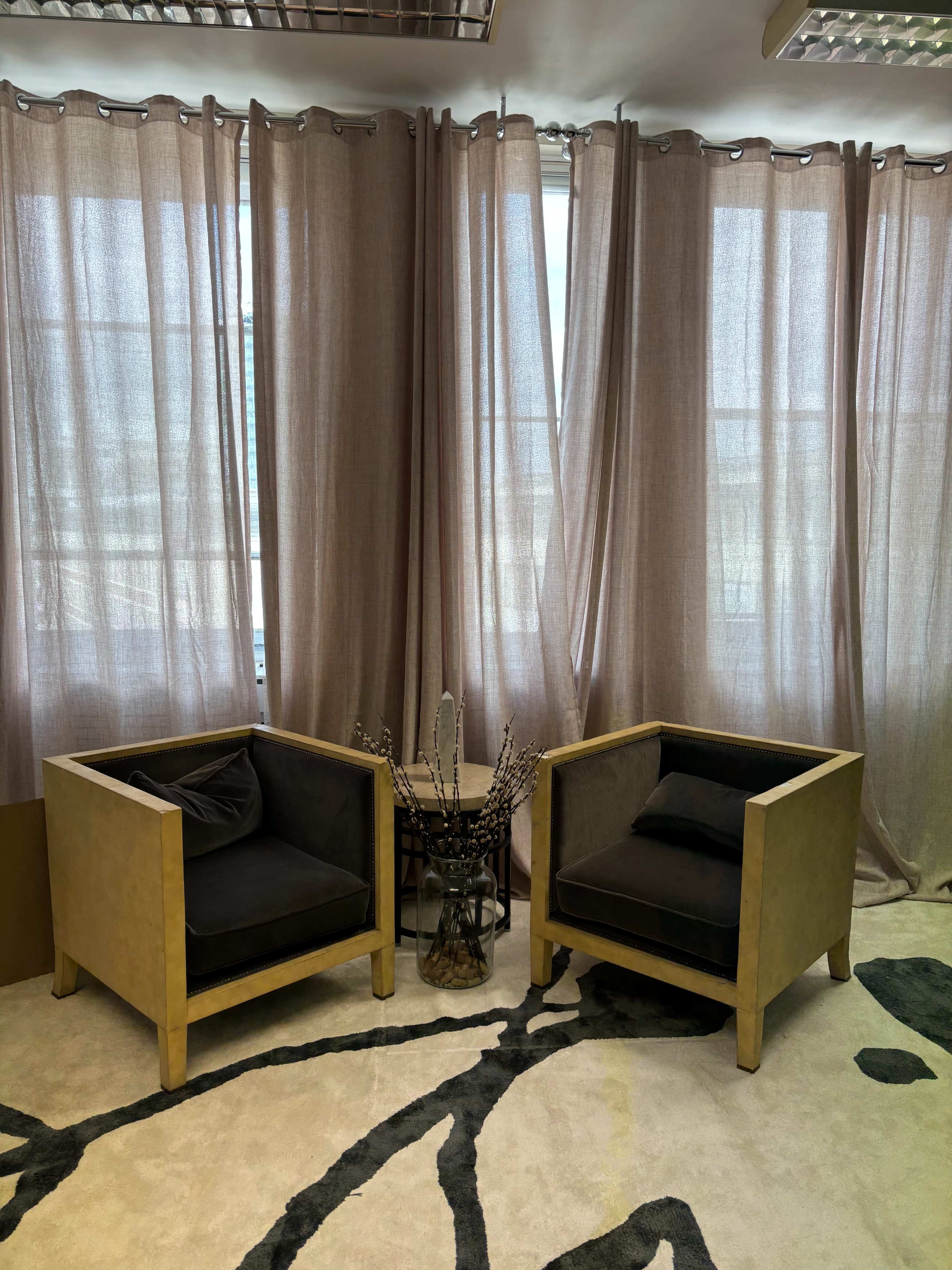 The image shows two modern armchairs positioned beside a decorative table with dried plants, set against sheer, light-colored curtains in a well-lit room.