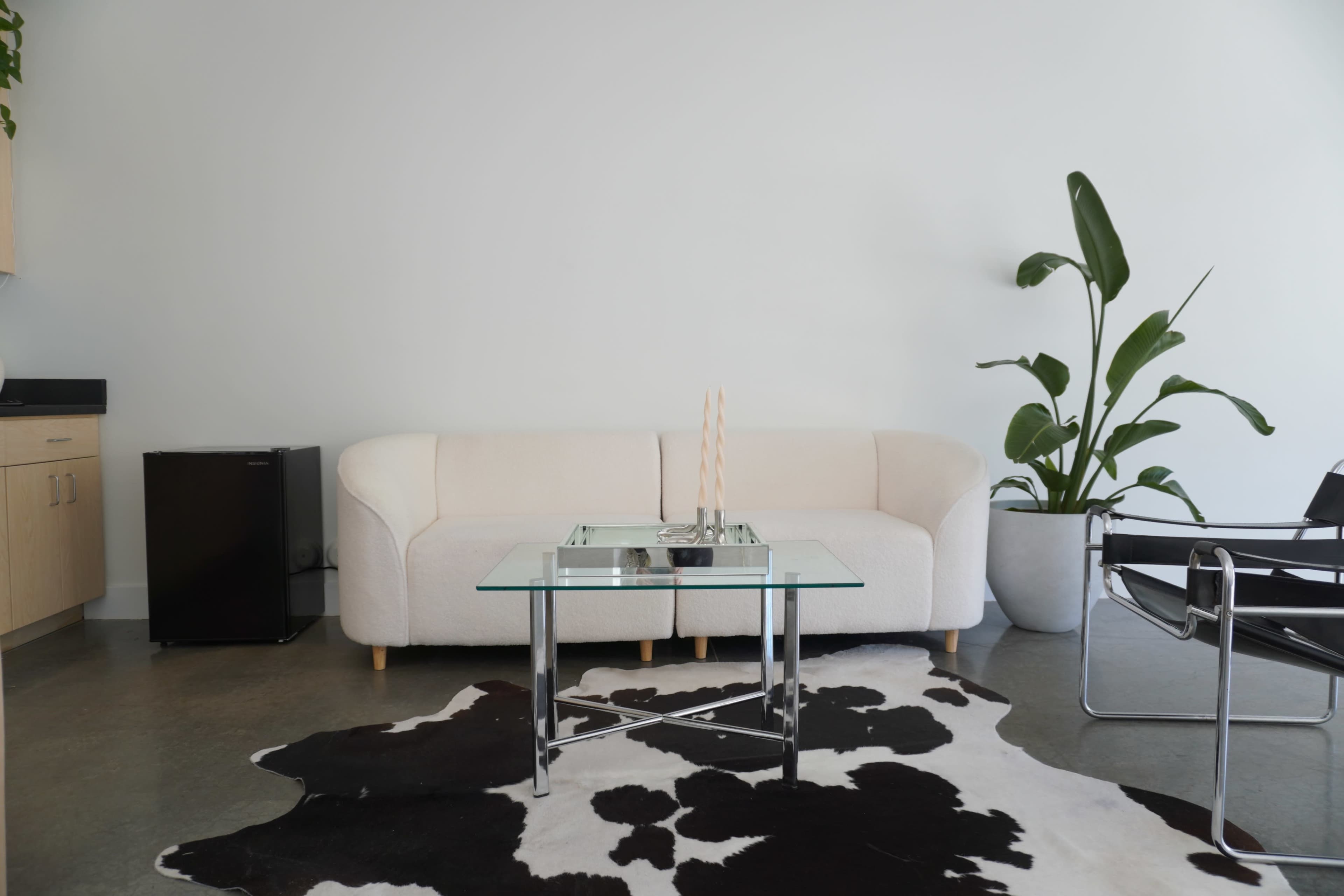 The image shows a modern living area with a white sofa, a glass coffee table, a black and white cowhide rug, and a potted plant.