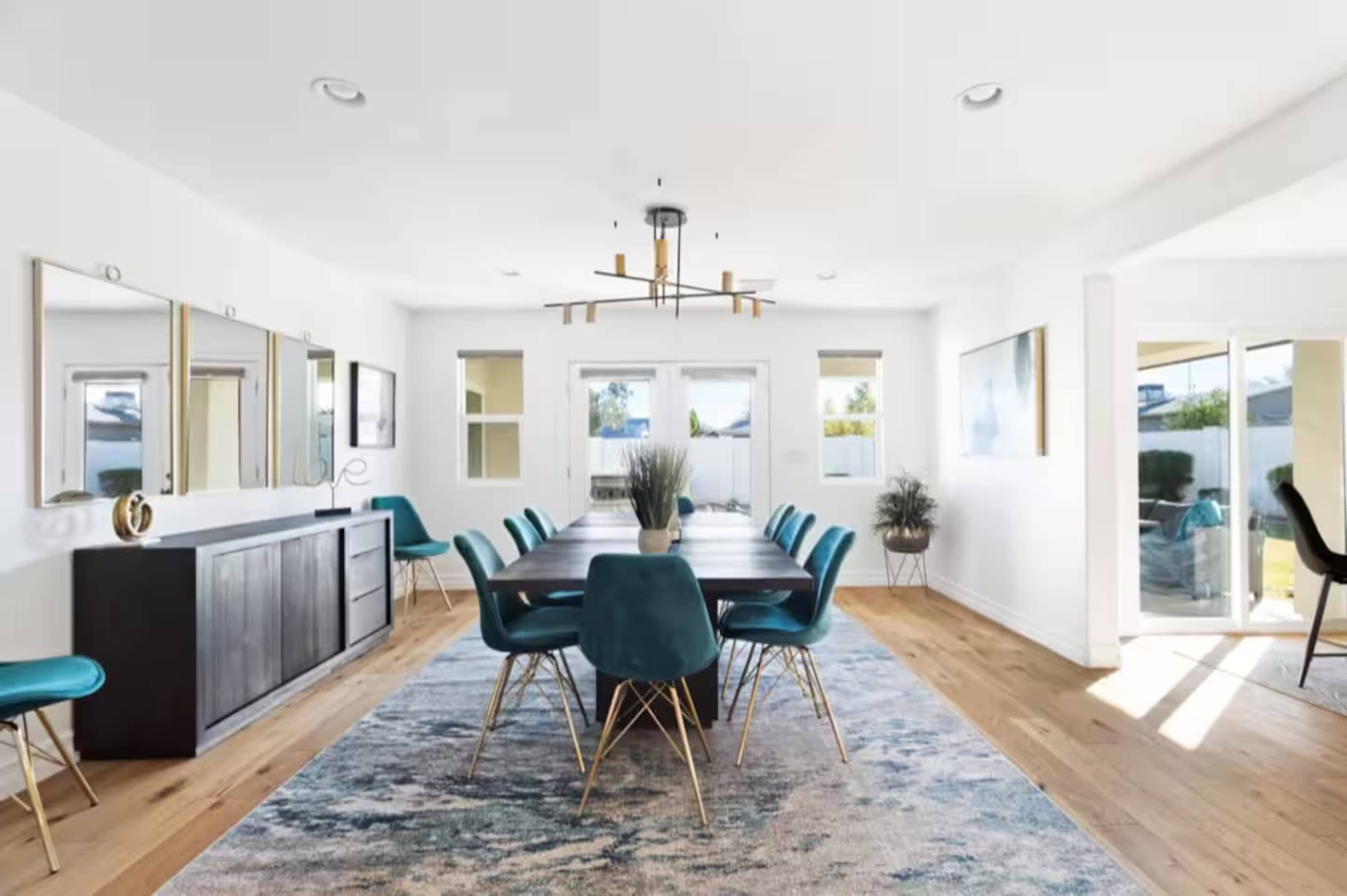 The image shows a modern dining room with a wooden table surrounded by teal chairs, a sideboard with mirrors, and large windows allowing natural light.