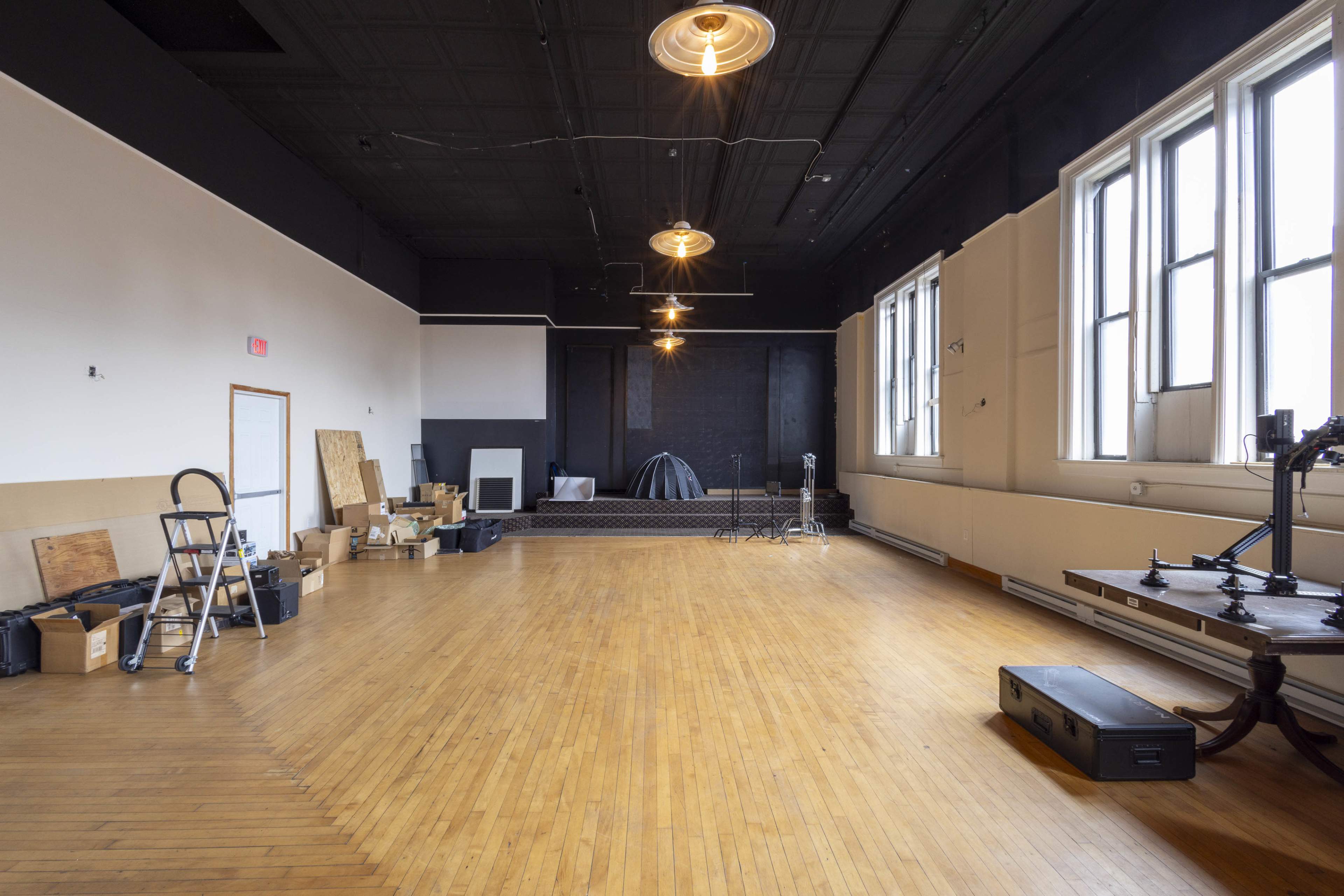 The image shows a spacious, empty room with hardwood flooring, large windows, and various pieces of equipment and boxes scattered around.