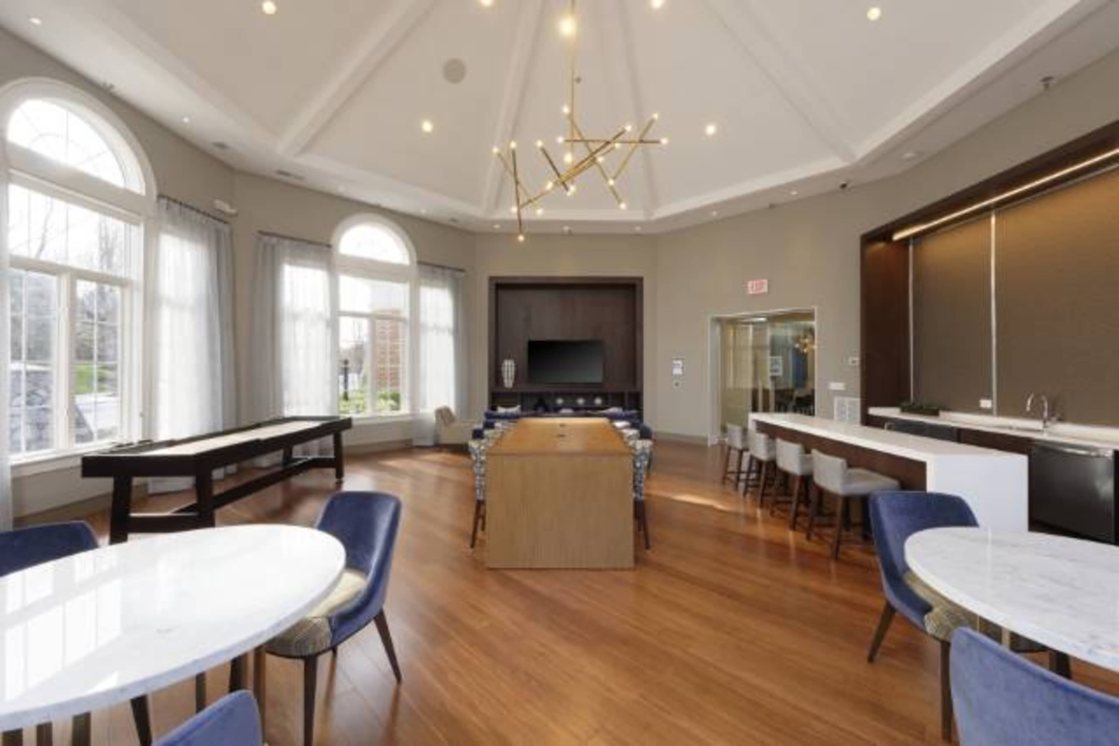 A spacious modern lounge area with large windows, a central dining table, a television on the wall, and a bar area along one side.