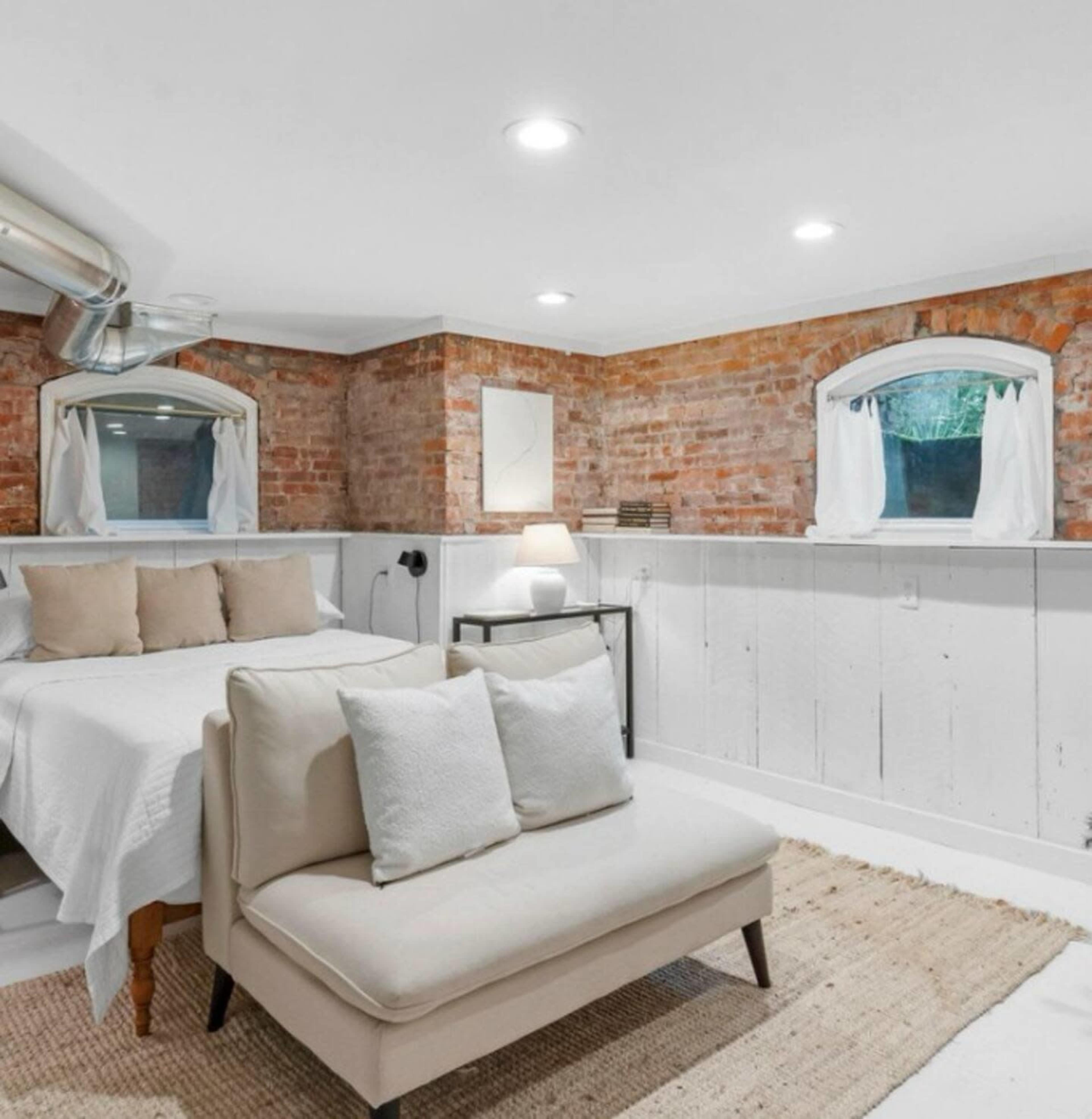 The image shows a bright basement room featuring a bed with neutral bedding, a small sofa, and exposed brick walls with white-paneled accents.