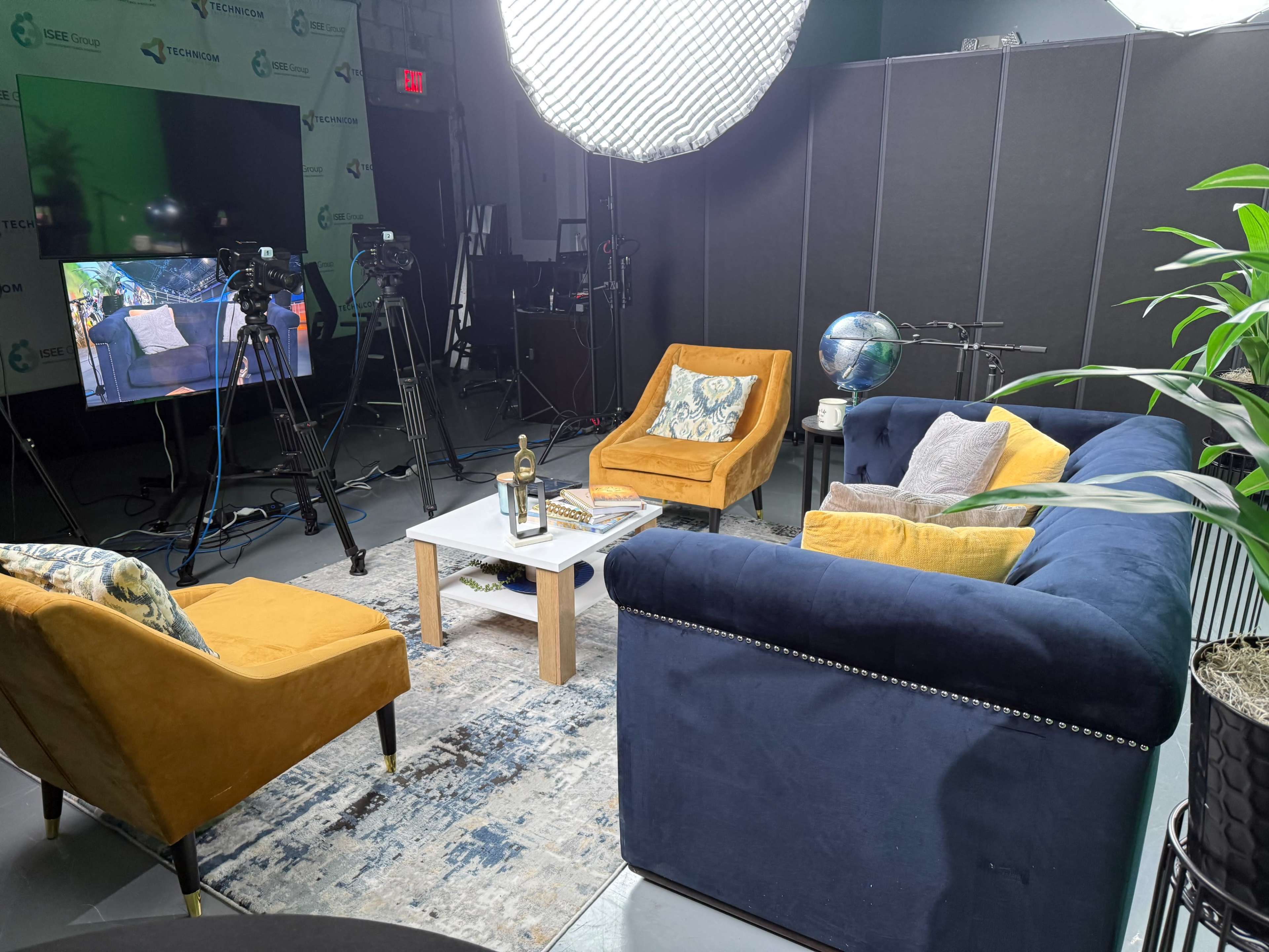 The image shows a modern studio setup with two yellow armchairs, a blue sofa, and a coffee table, surrounded by cameras and lighting equipment.
