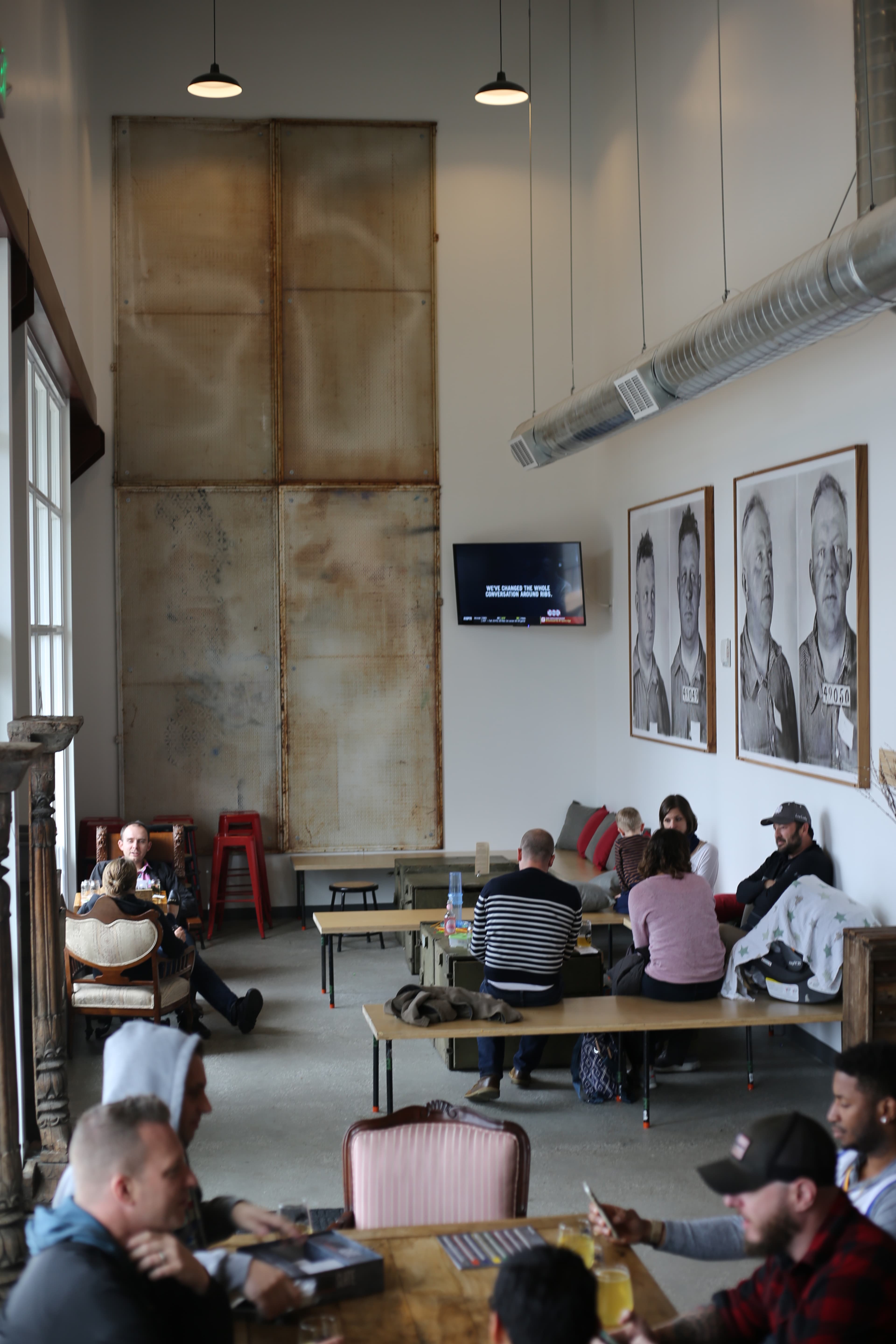 The image shows a spacious café with a mix of patrons seated at wooden tables, large portraits on the walls, and a television mounted above.