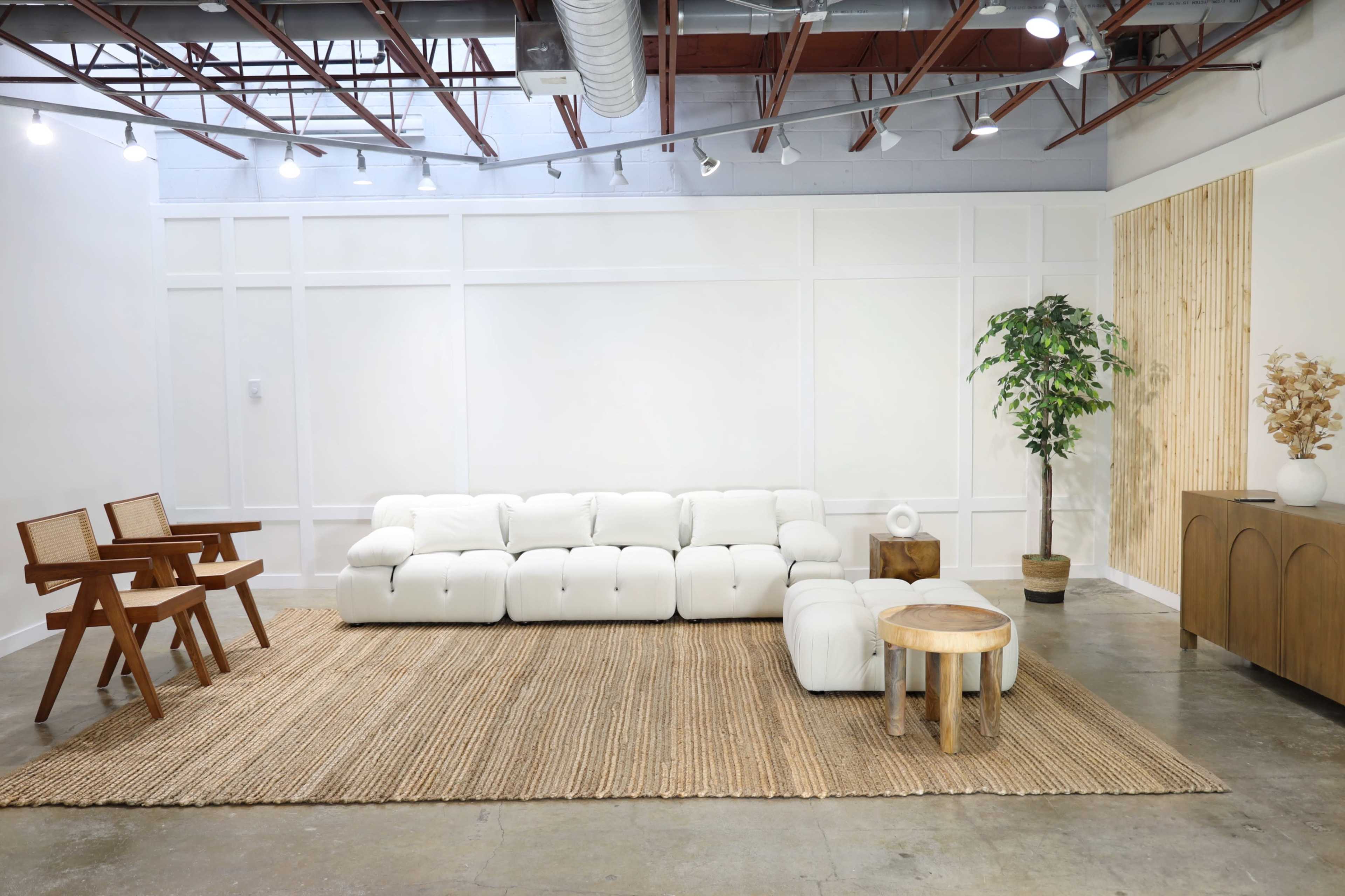 A modern living room with a white sectional sofa, a woven rug, a wooden side table, and a potted plant against a white wall.