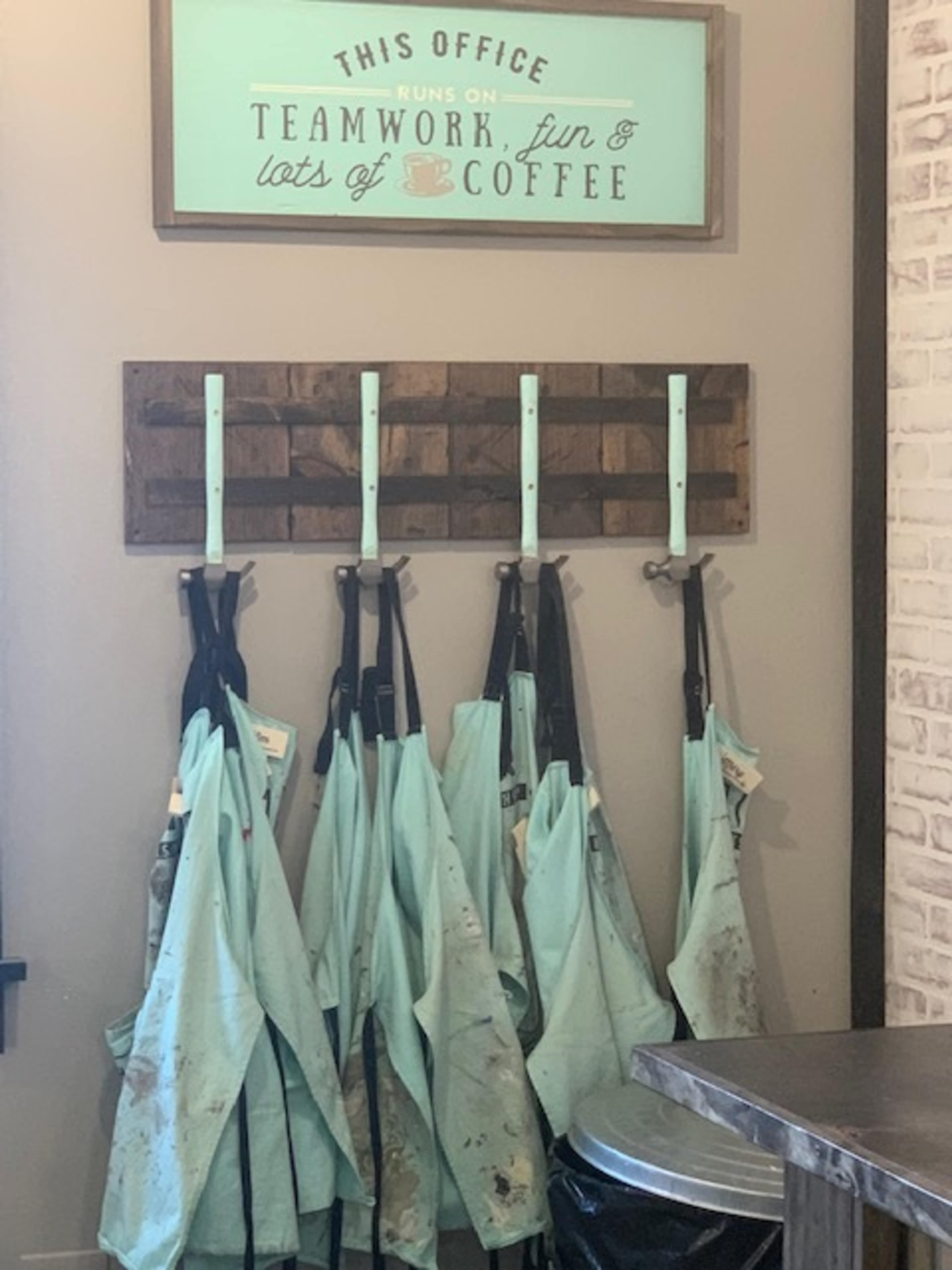 A wall in an office with a sign that says "This office runs on teamwork, fun, and lots of coffee" above a rack holding several light blue, dirty aprons.