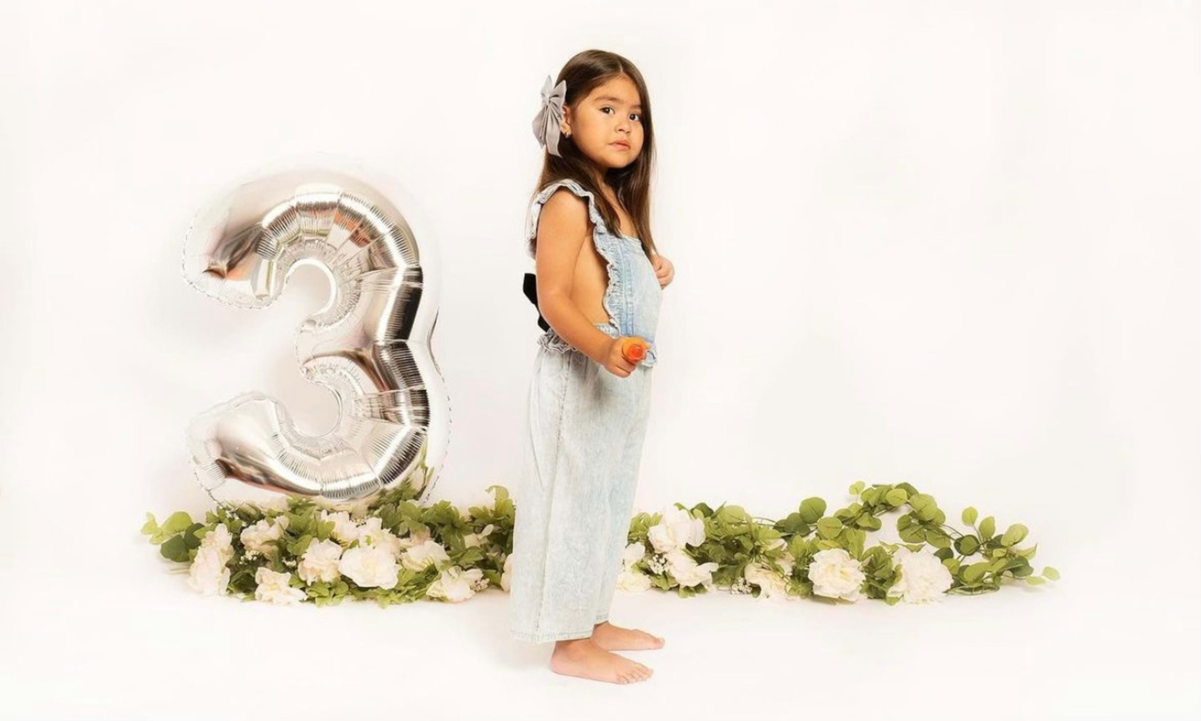 A girl stands next to a large silver balloon shaped like the number three, with flowers and greenery arranged on the floor.