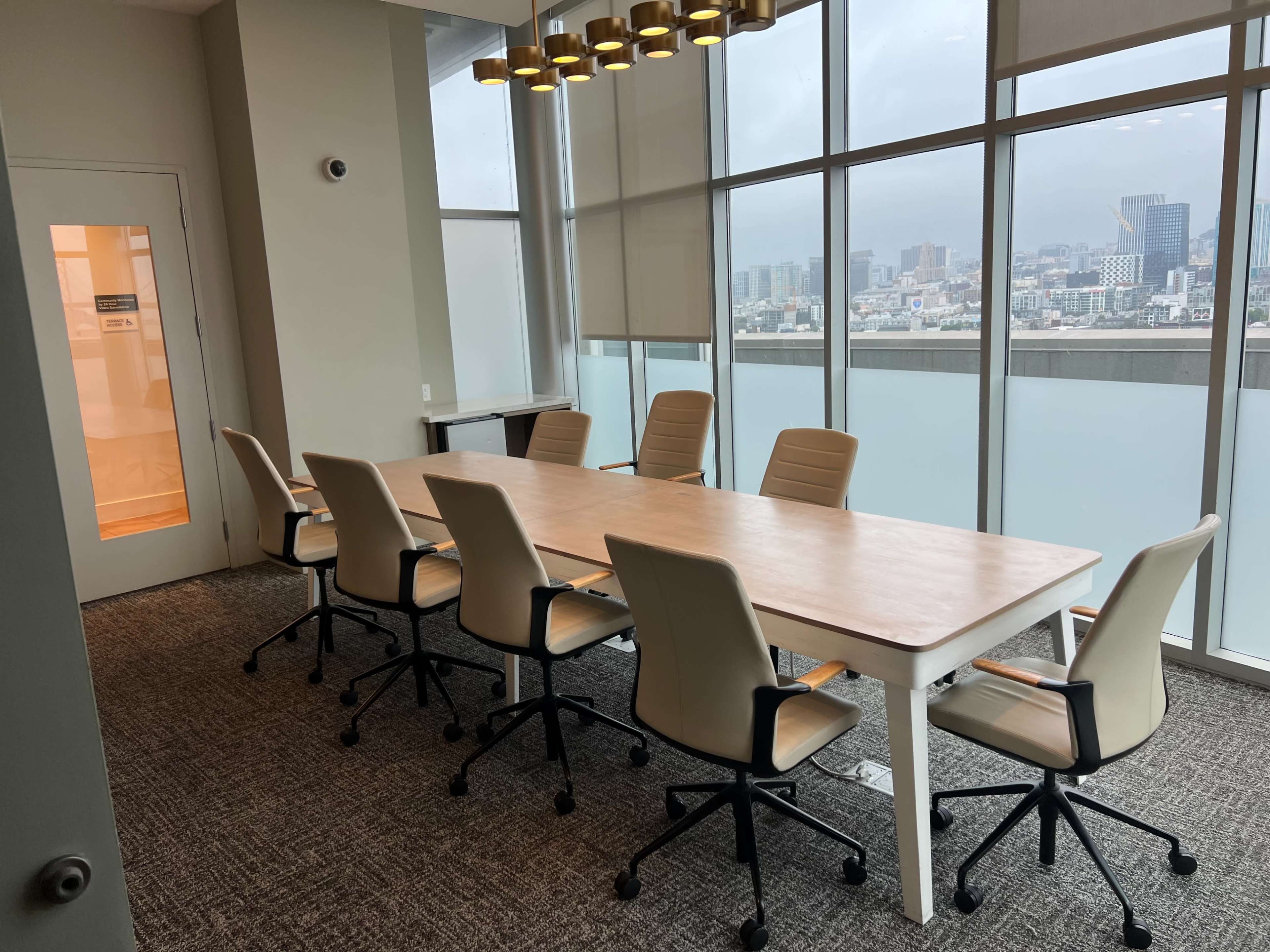 A conference room features a long wooden table surrounded by six beige rolling chairs, with large windows showcasing a city skyline in the background.