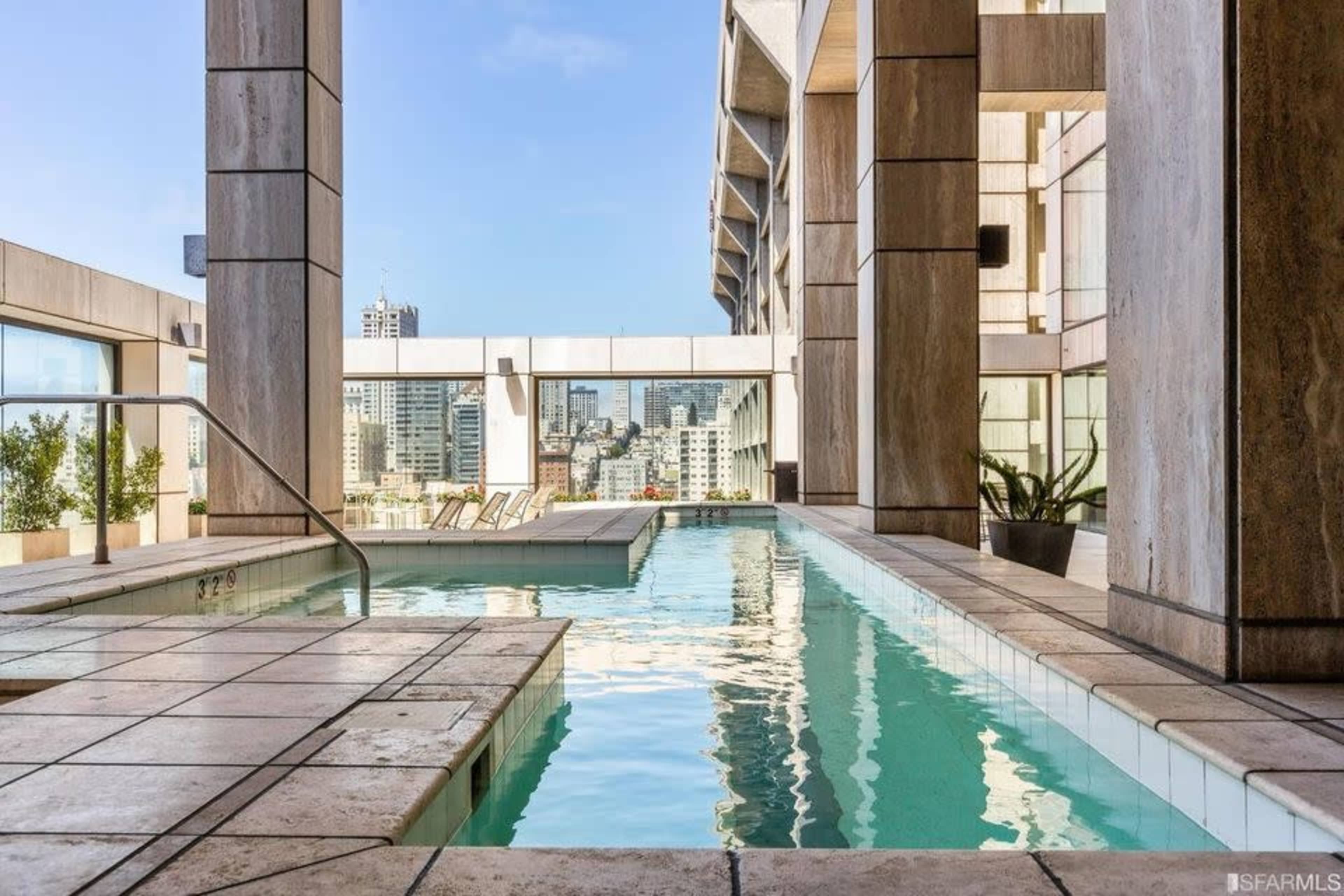 A modern rooftop swimming pool surrounded by concrete pillars and a view of a city skyline in the background.