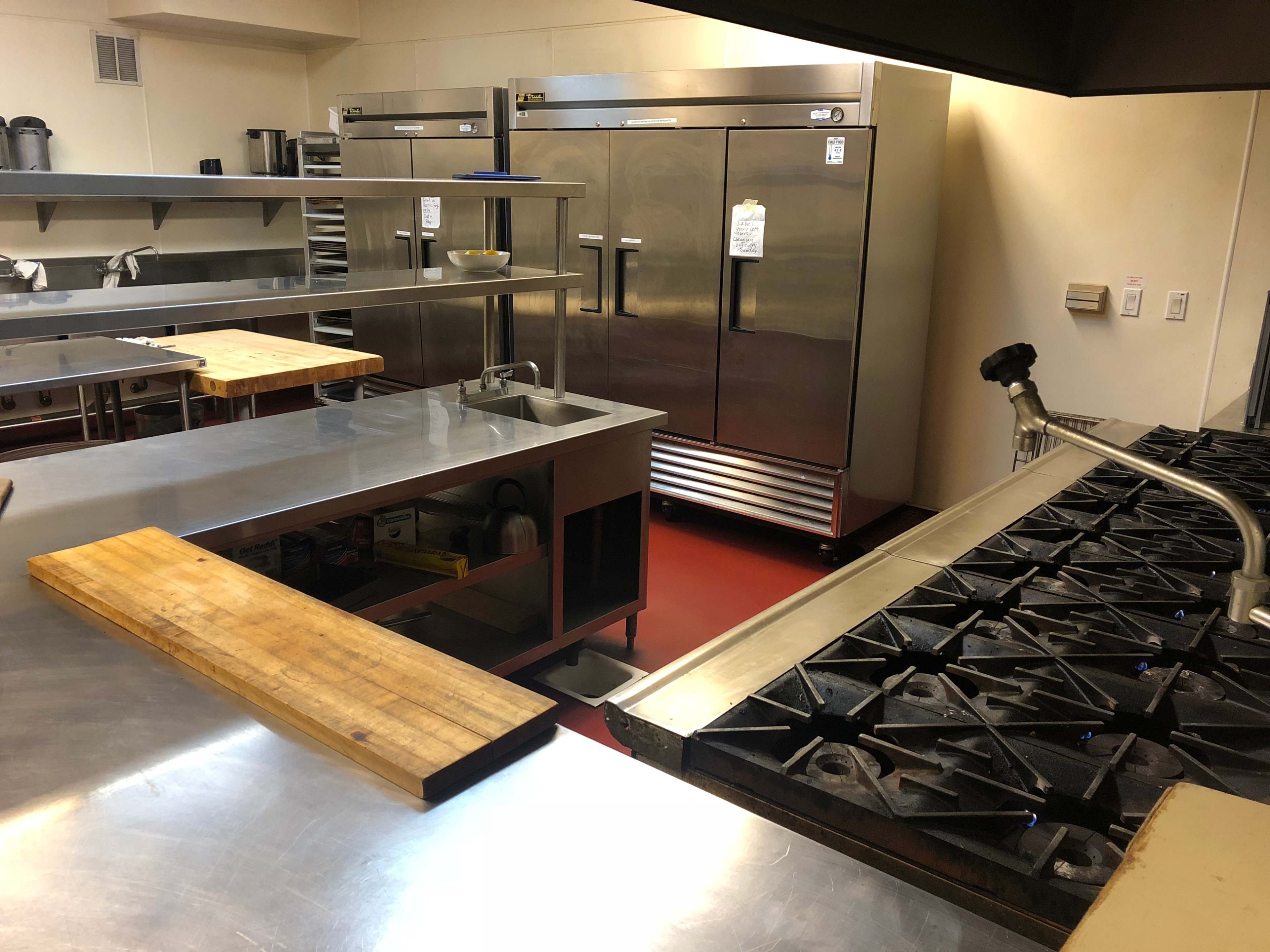 A commercial kitchen with stainless steel appliances, a wooden cutting board, and a gas range with multiple burners.
