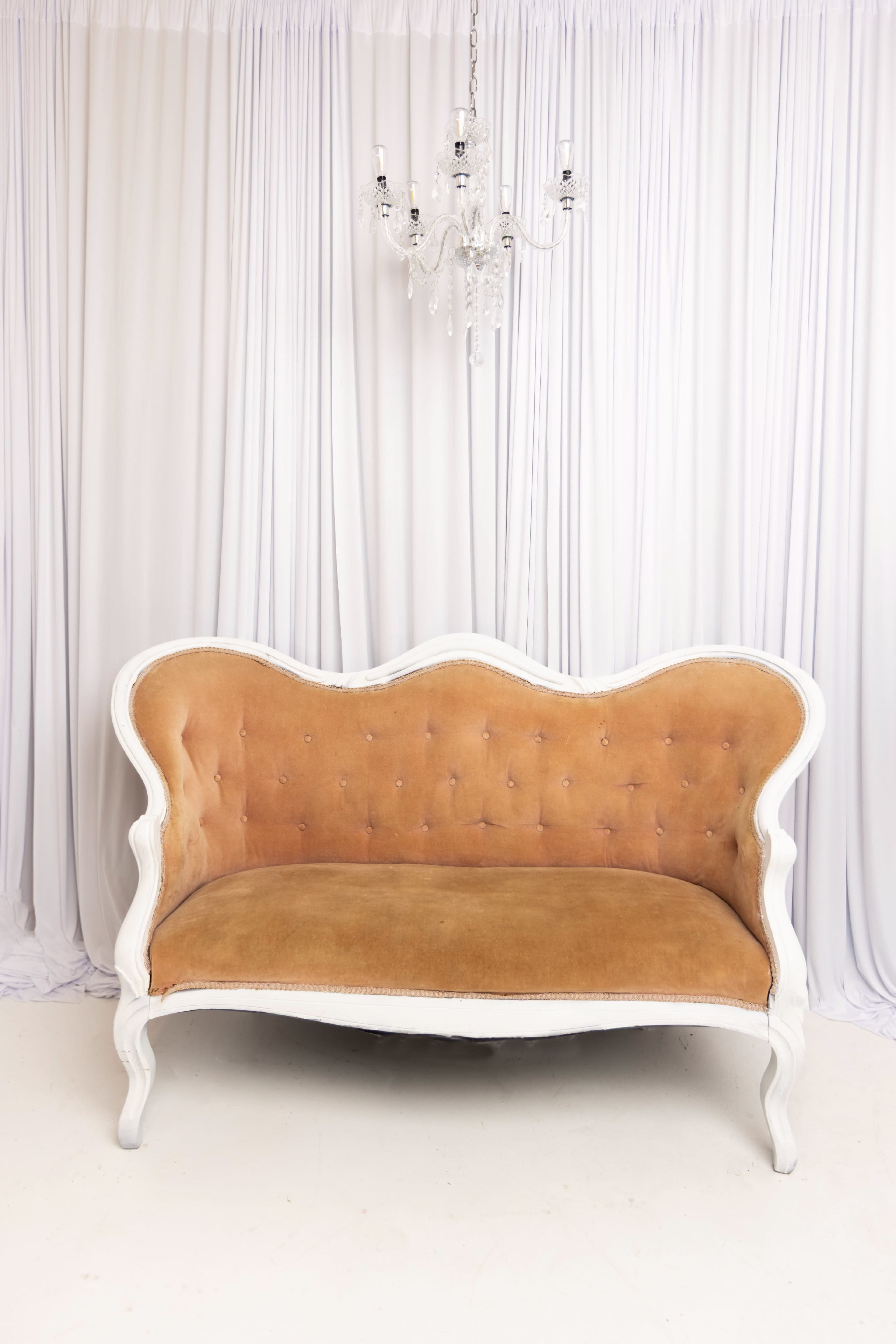 A vintage-style, tufted loveseat with a light tan upholstery is positioned in front of a white draped backdrop, under a crystal chandelier.