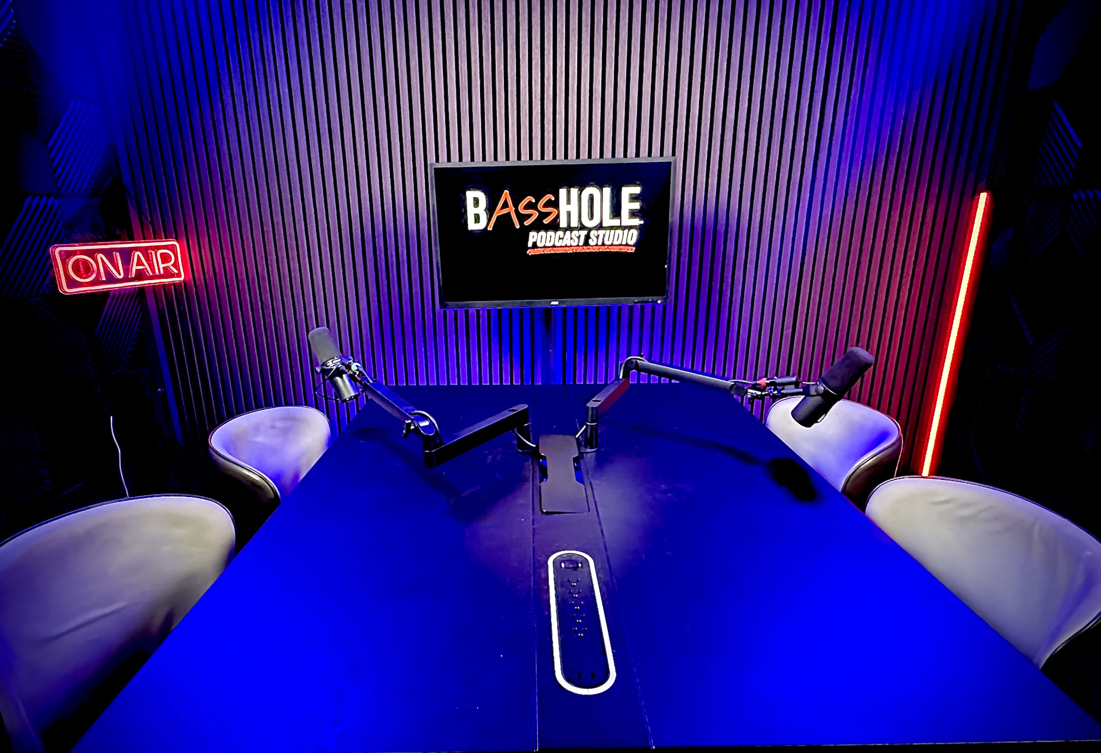 The image shows a podcast studio setup with a prominent sign reading "BASSHOLE PODCAST STUDIO" displayed on a screen, two microphones positioned on a table, and ambient lighting in purple and blue tones.