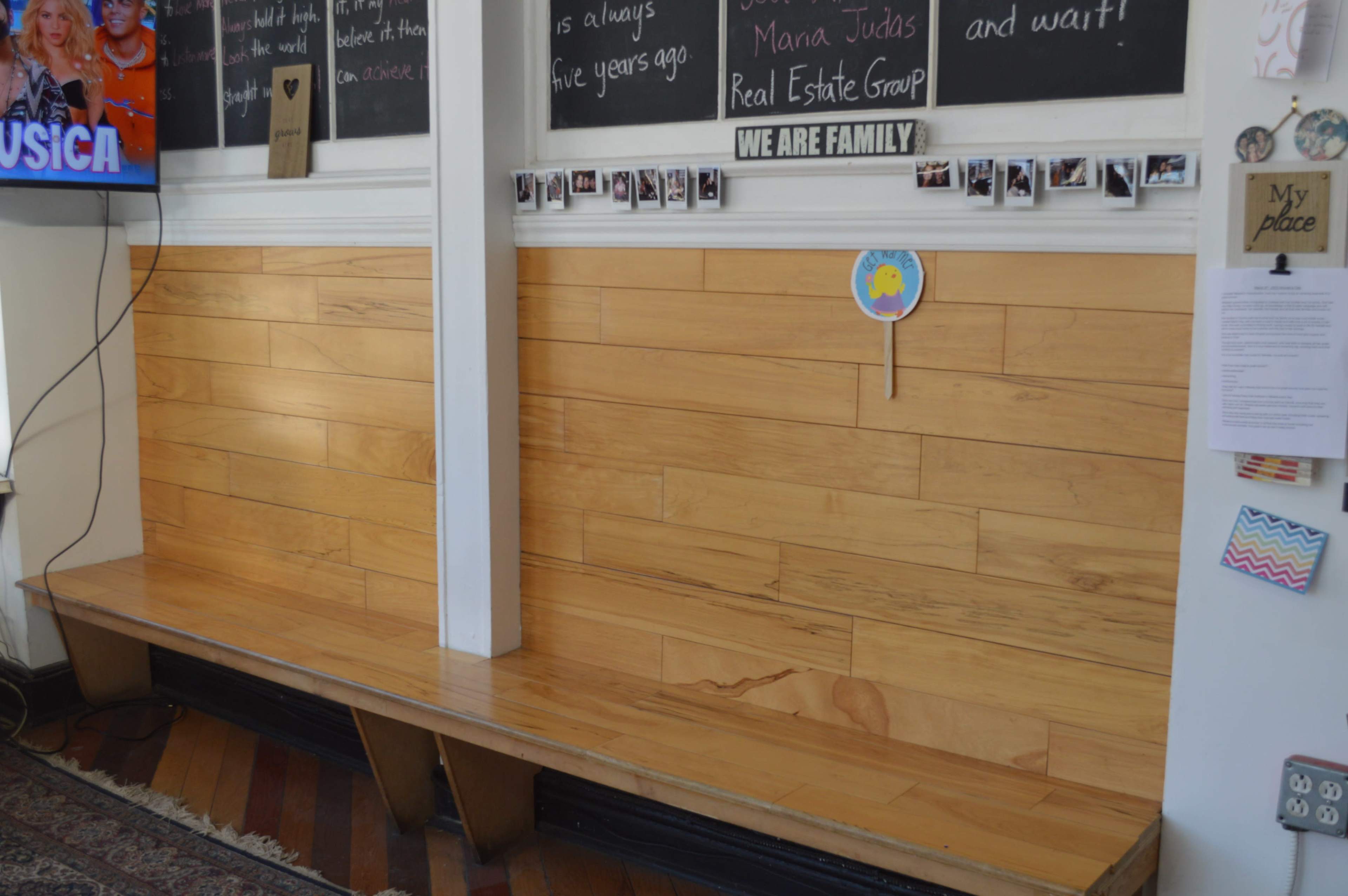 A wooden bench against a wall with a television mounted nearby and a bulletin board covered in photographs and notes.