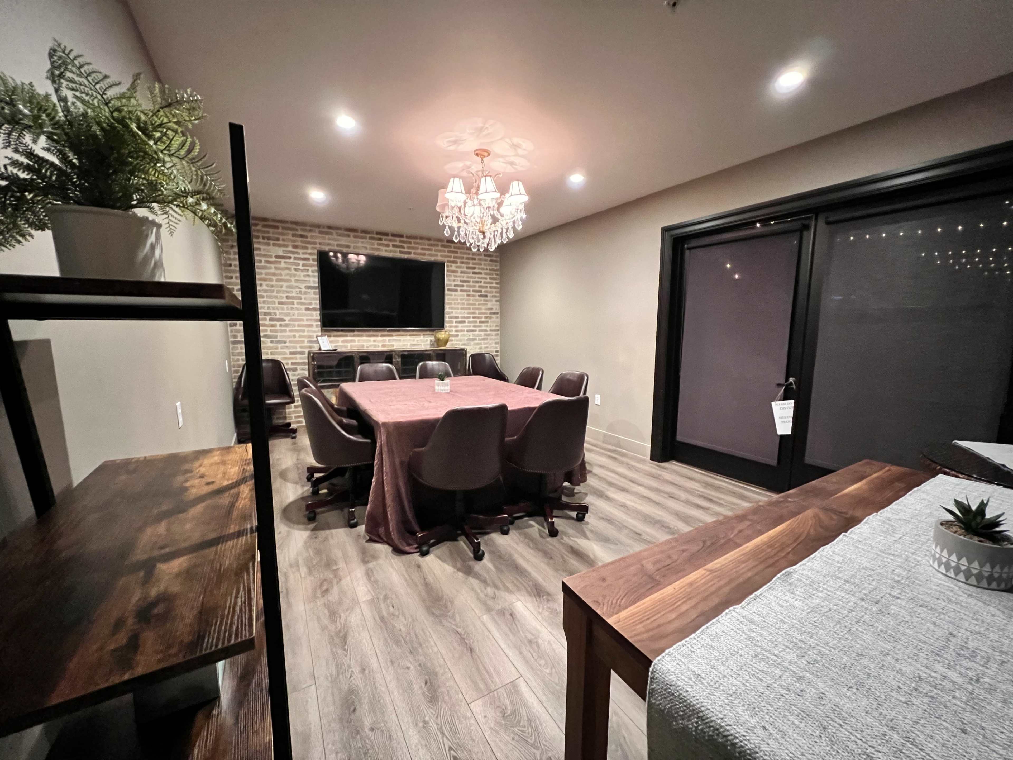 A conference room features a large table surrounded by chairs, with a TV on a brick wall and a chandelier overhead.