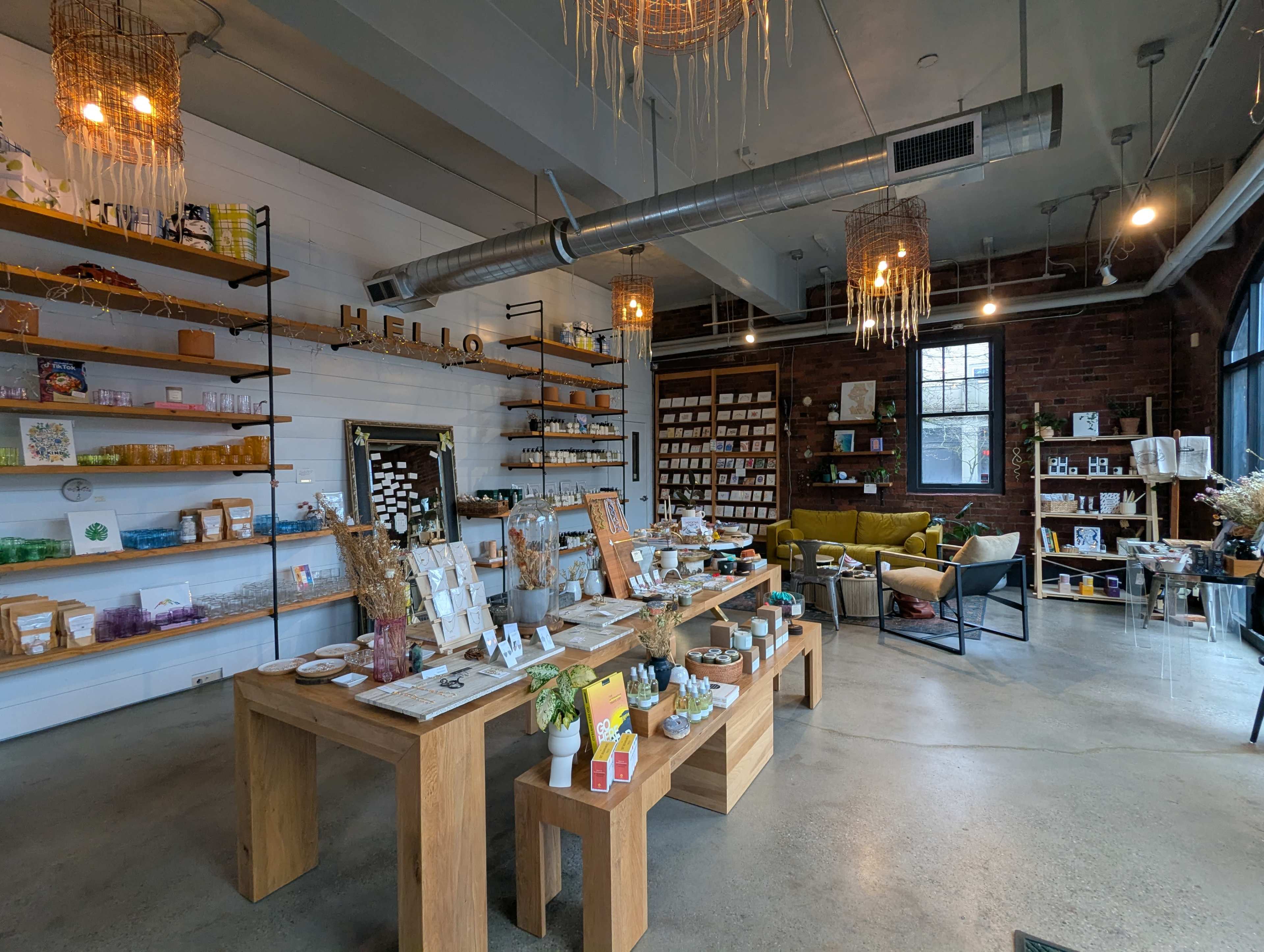 The image shows a modern retail space featuring wooden display tables, shelves stocked with various products, and a cozy seating area.