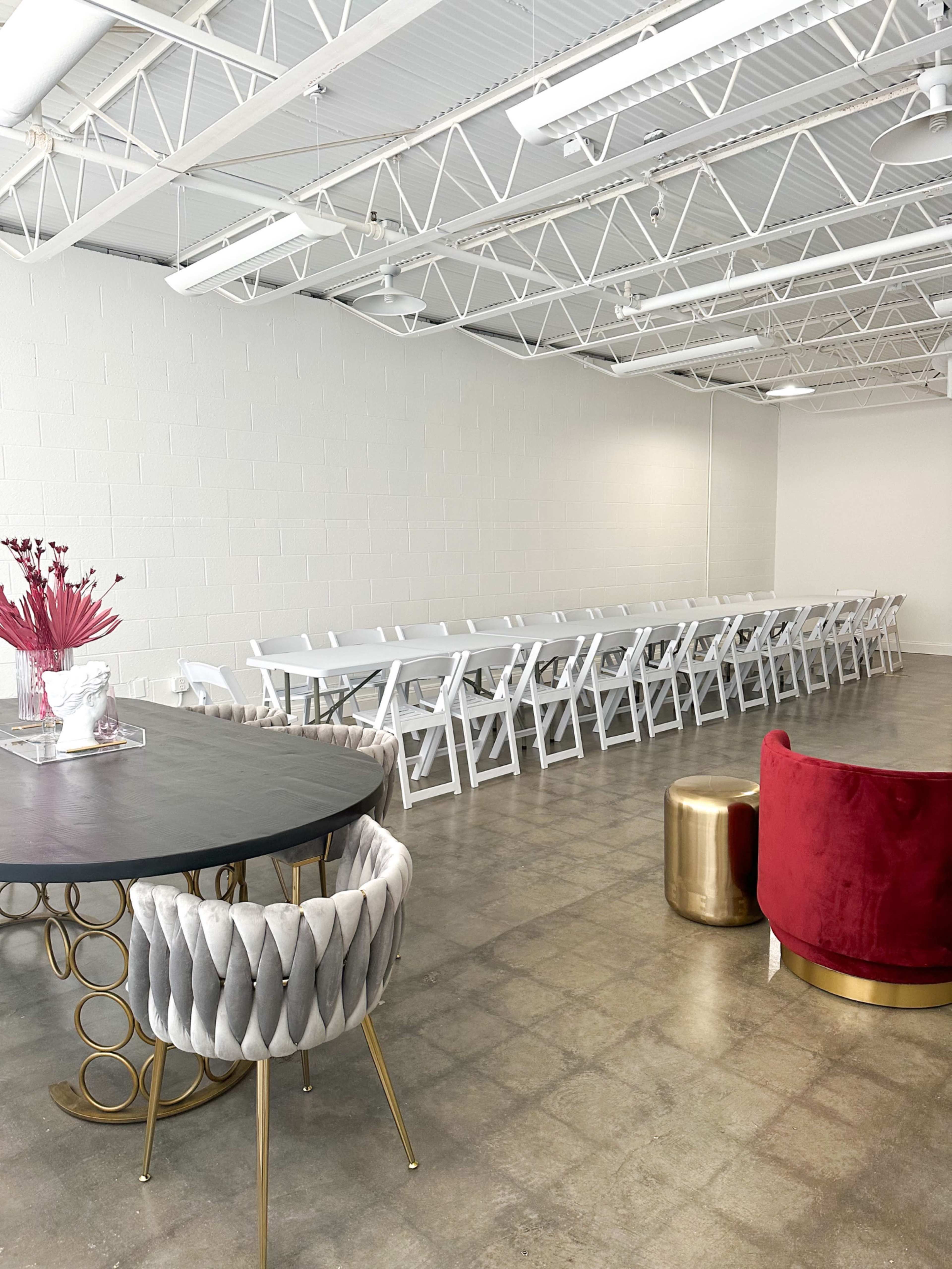 The image shows a spacious indoor venue featuring a long table lined with white folding chairs, a round table with a decorative centerpiece, and a red upholstered chair.