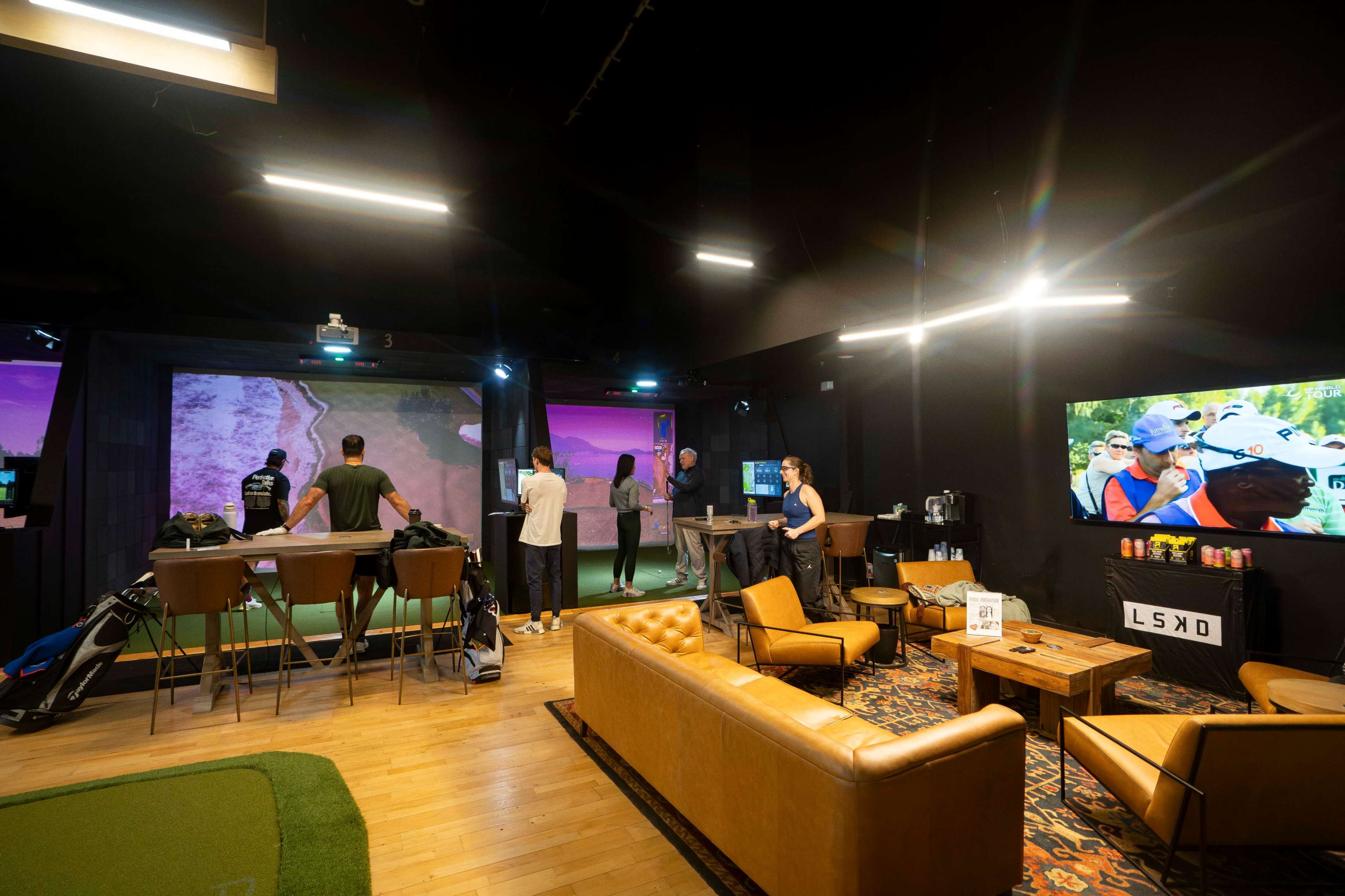 The image shows a modern indoor golf simulation facility with people using golf clubs in front of large screens displaying golf courses.