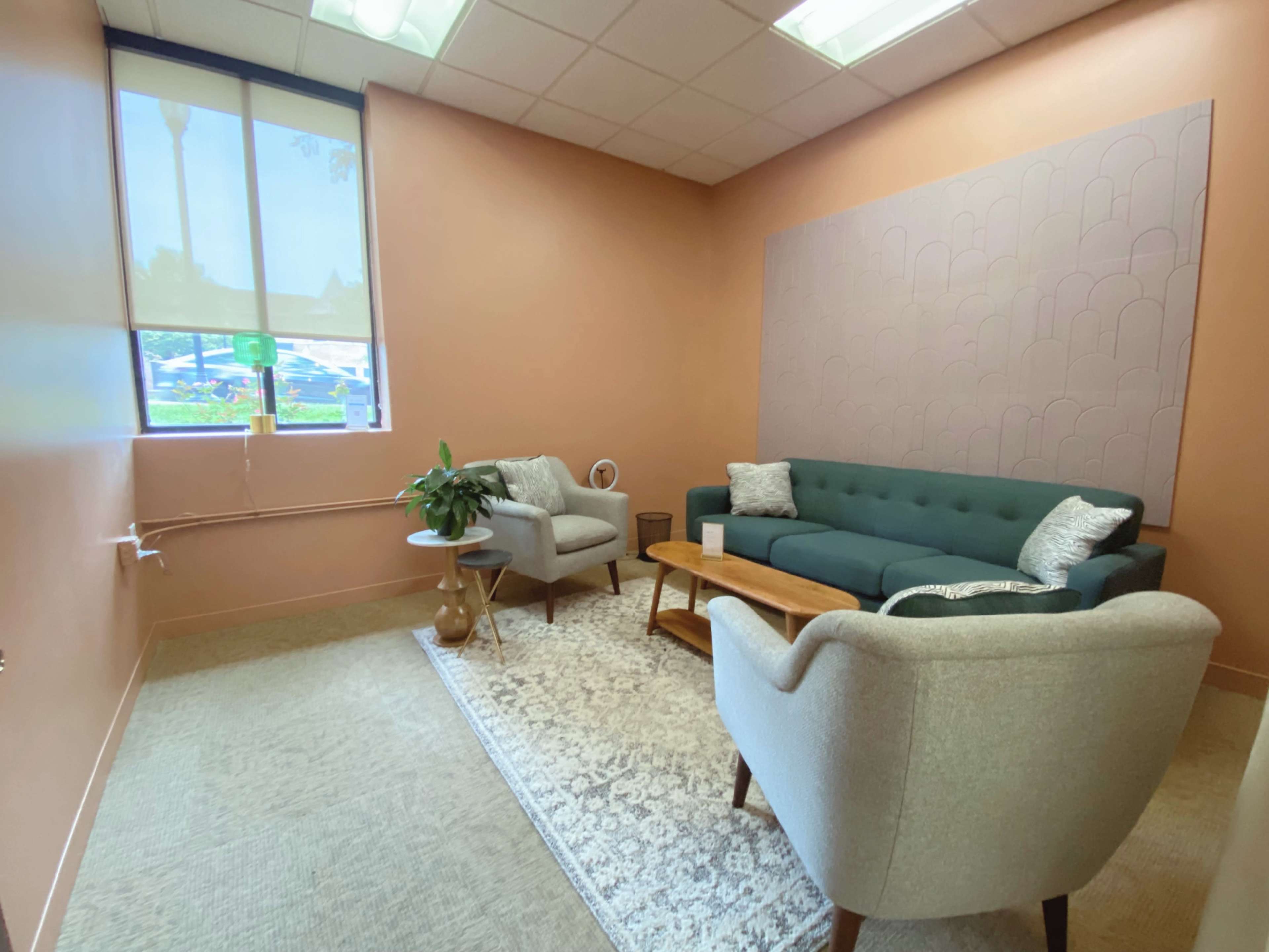 The image shows a small, well-lit lounge with a blue sofa, two light gray armchairs, a wooden coffee table, and a decorative rug.
