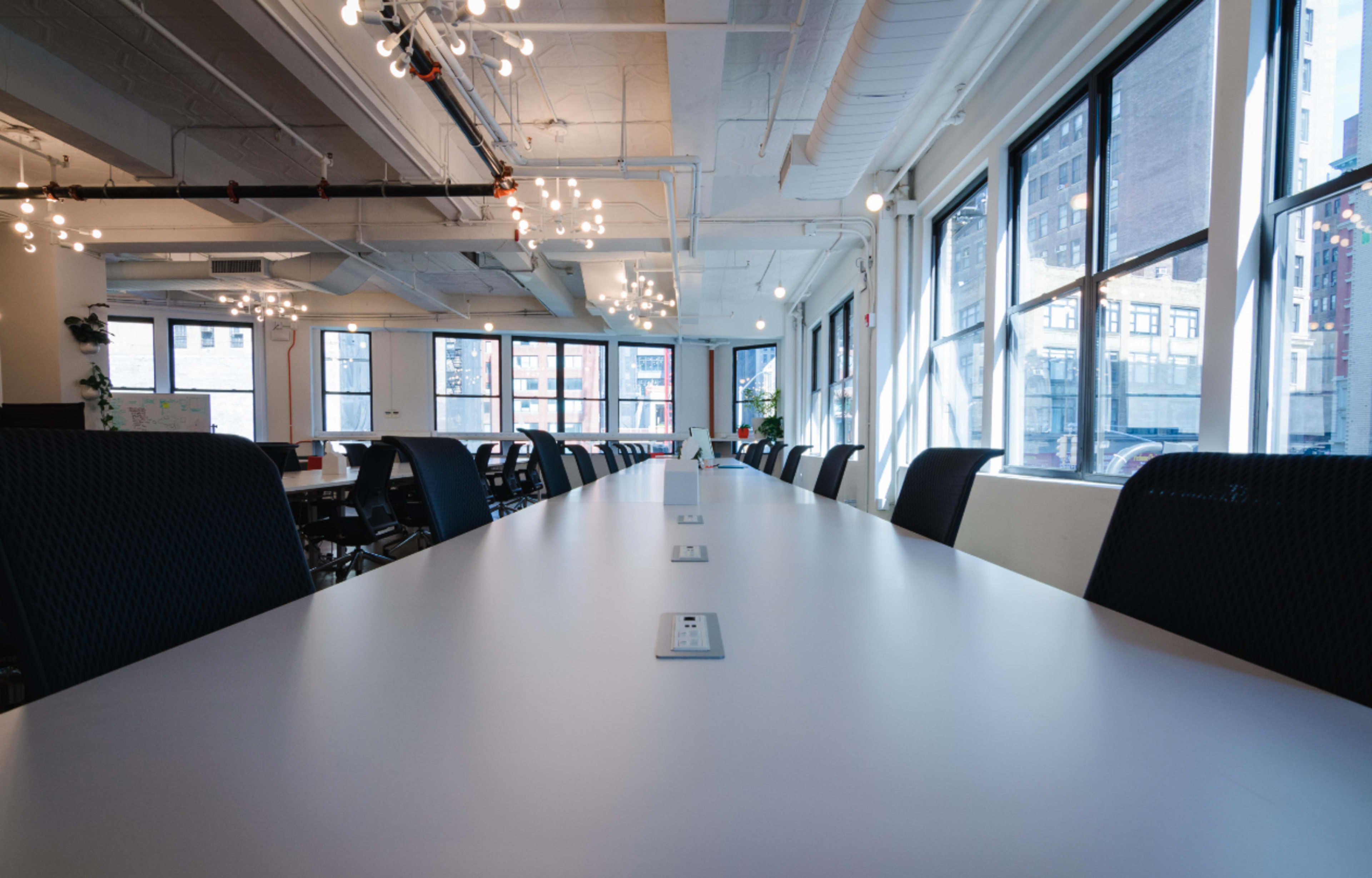A long conference table with black chairs is set in a brightly lit office space featuring large windows.