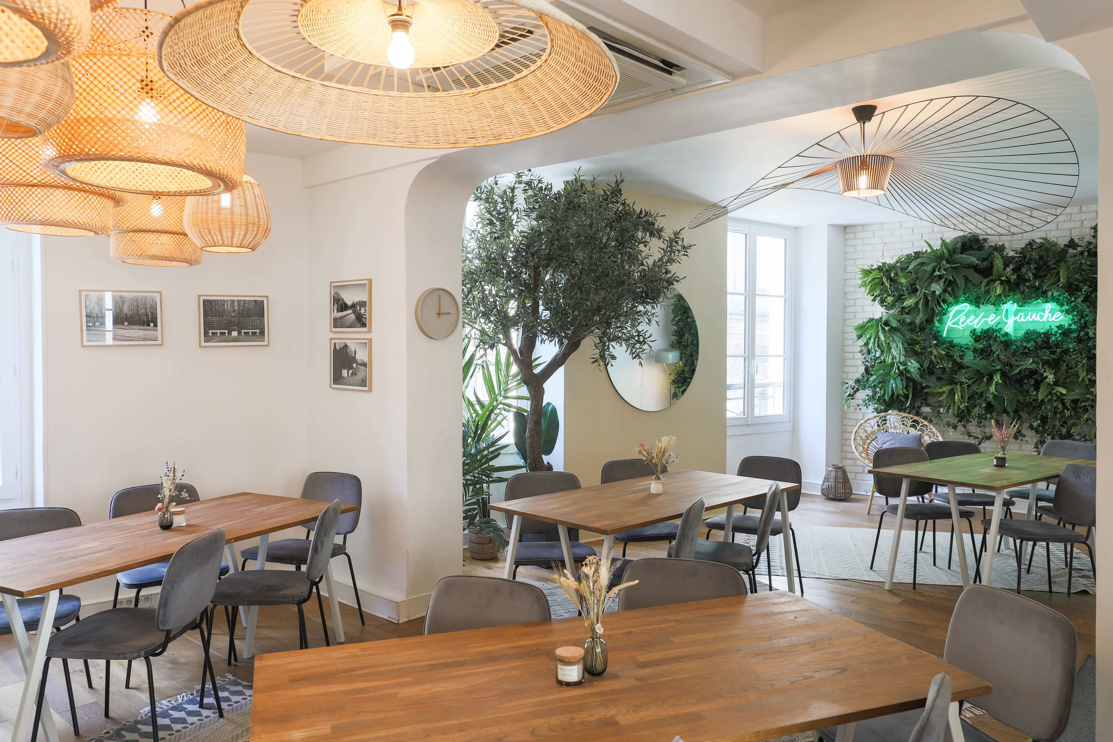 A modern restaurant features wooden tables, woven light fixtures, green plants, and a neon sign against a stylish backdrop.