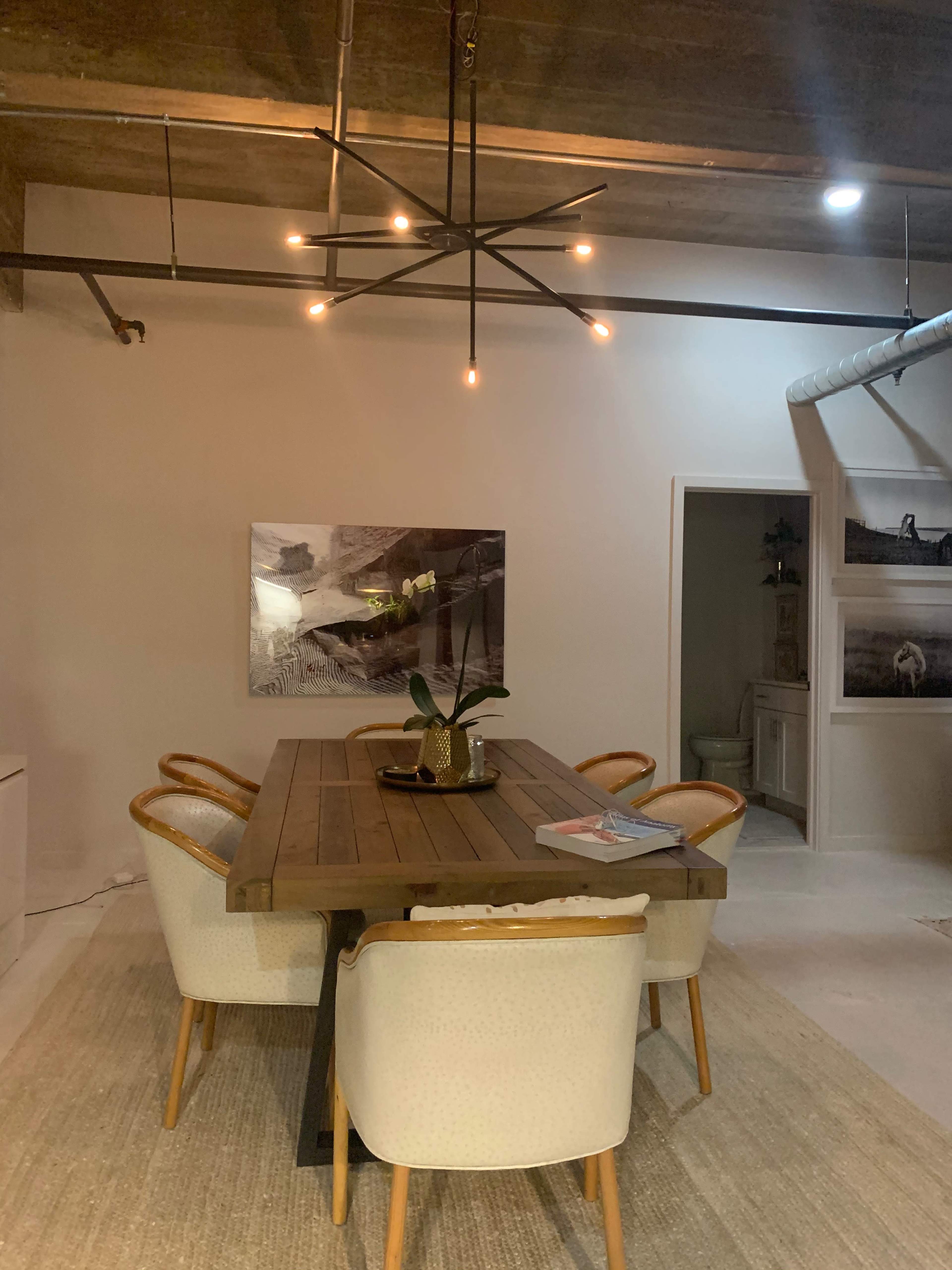 The image shows a dining area featuring a wooden table surrounded by upholstered chairs, illuminated by a modern chandelier, with artwork on the walls and a doorway leading to another room.