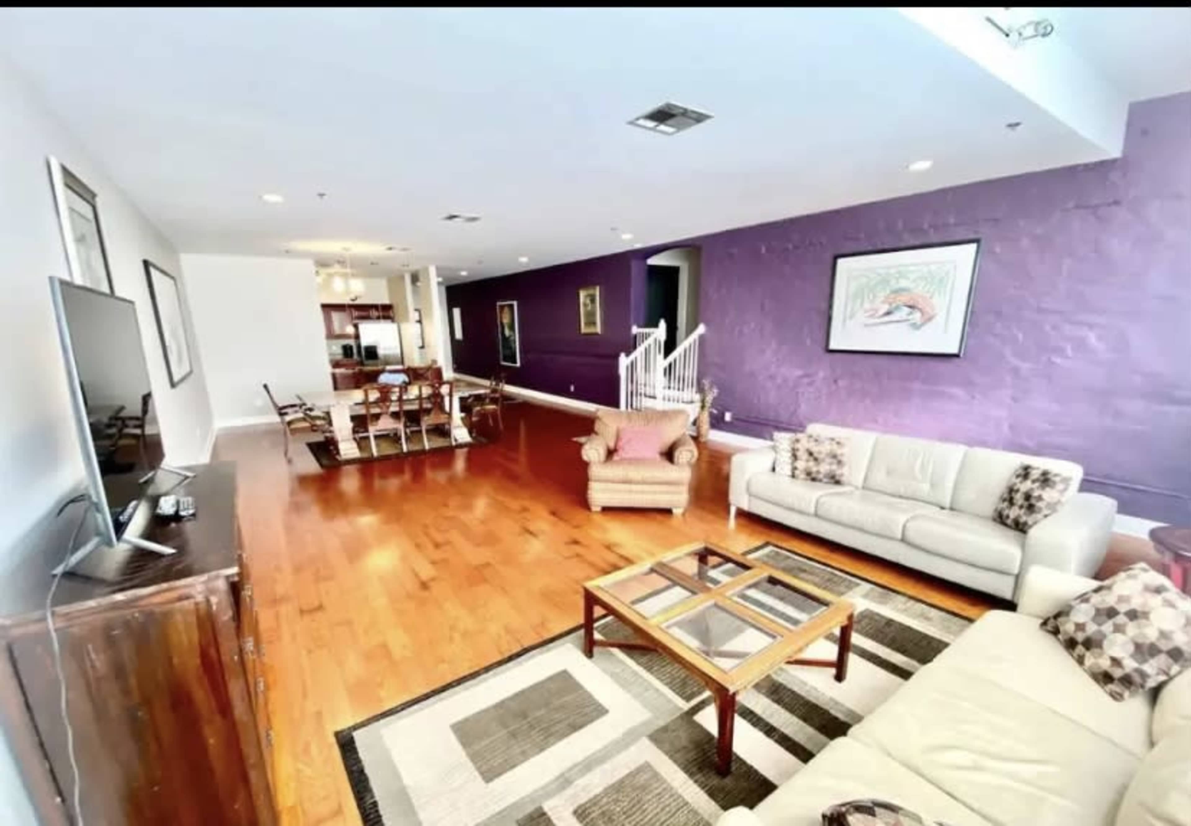 A spacious living room features hardwood floors, a purple accent wall, and a seating area with two sofas and a coffee table, leading to a dining area and kitchen.