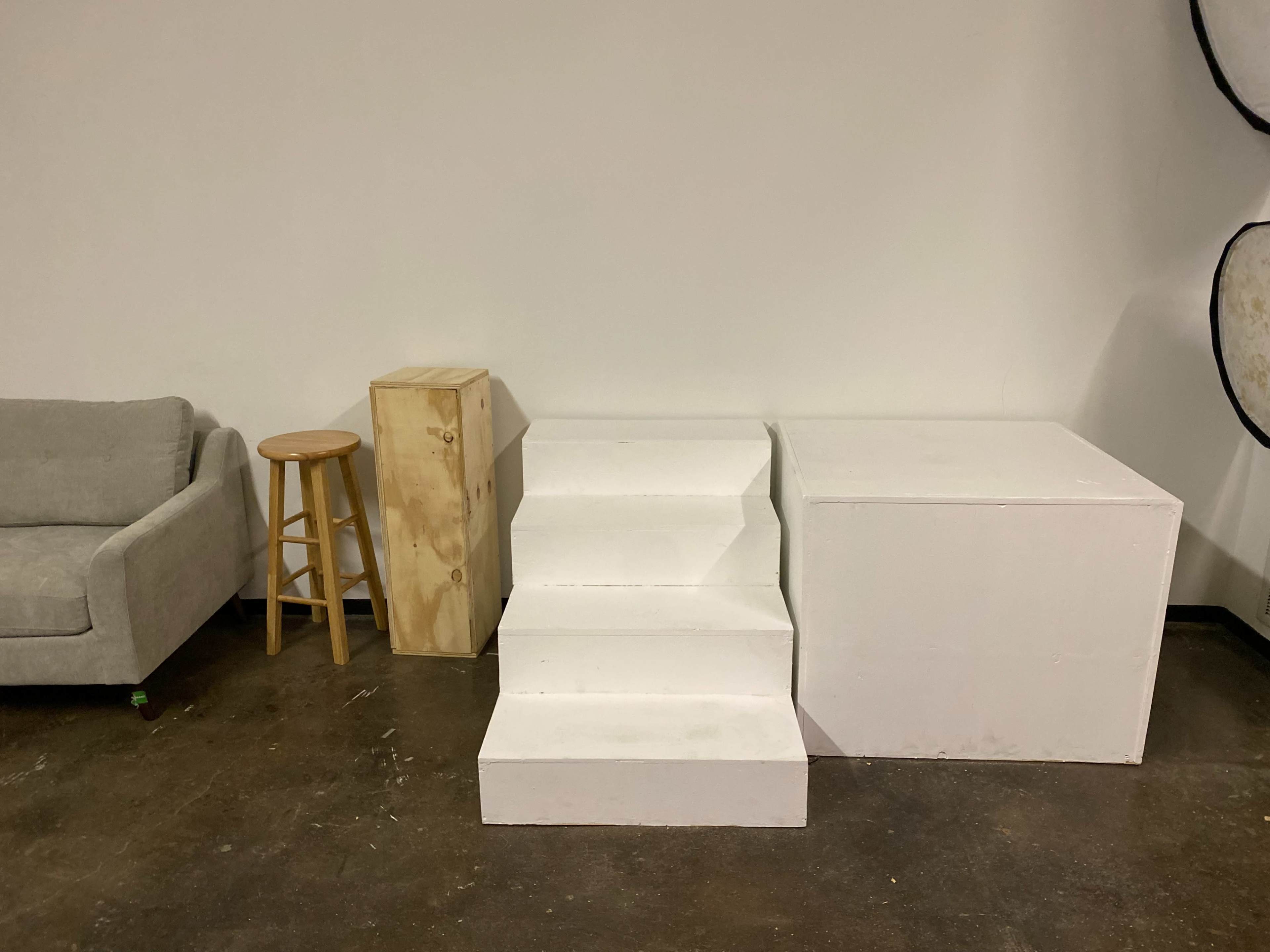 A simple interior scene featuring a gray couch, a wooden stool, a stack of wooden boxes, three white steps, and a large white cube against a plain wall.
