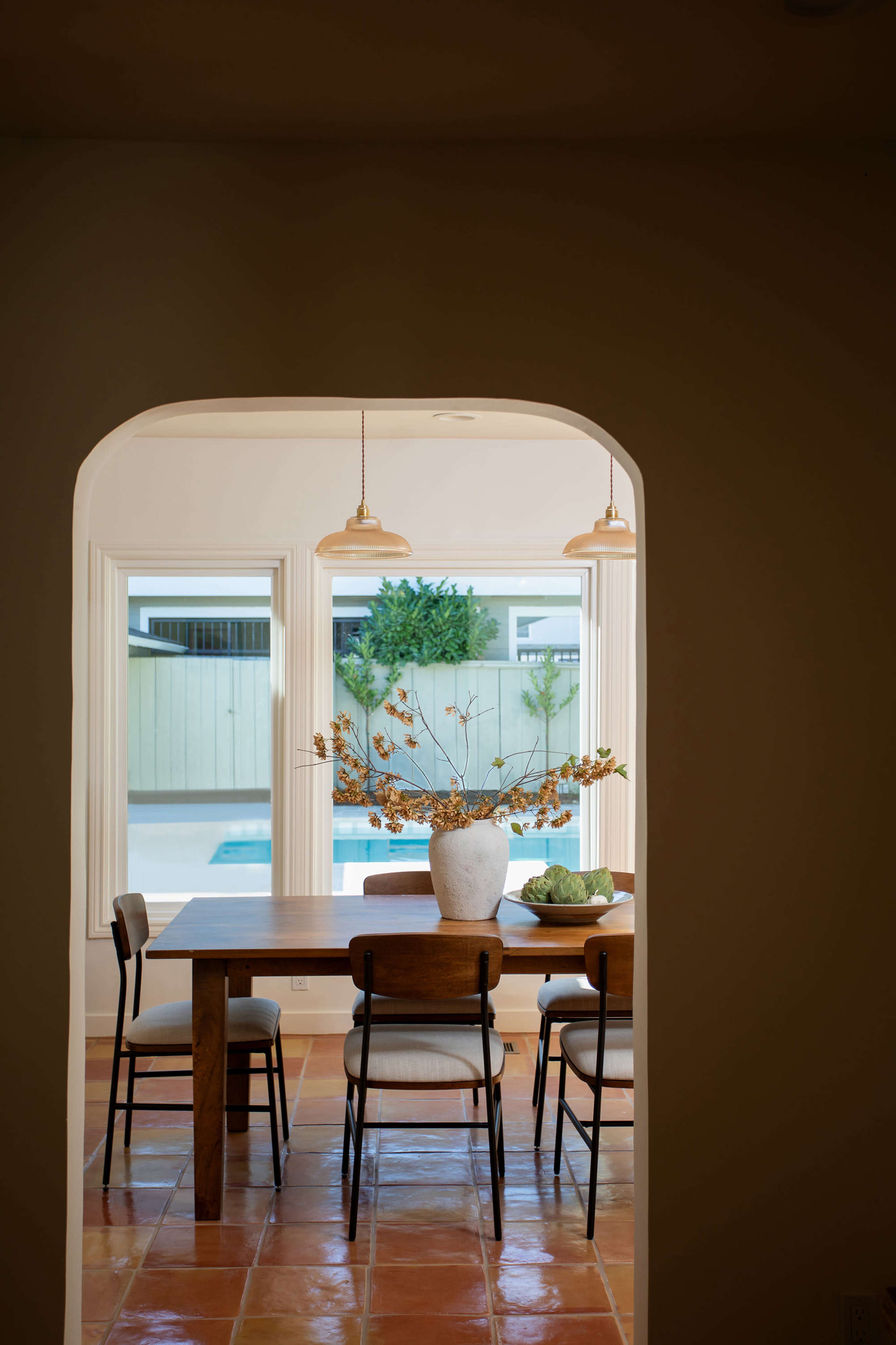 A dining room with a wooden table and chairs is framed by an archway, featuring a large vase of branches and a view of a pool outside.