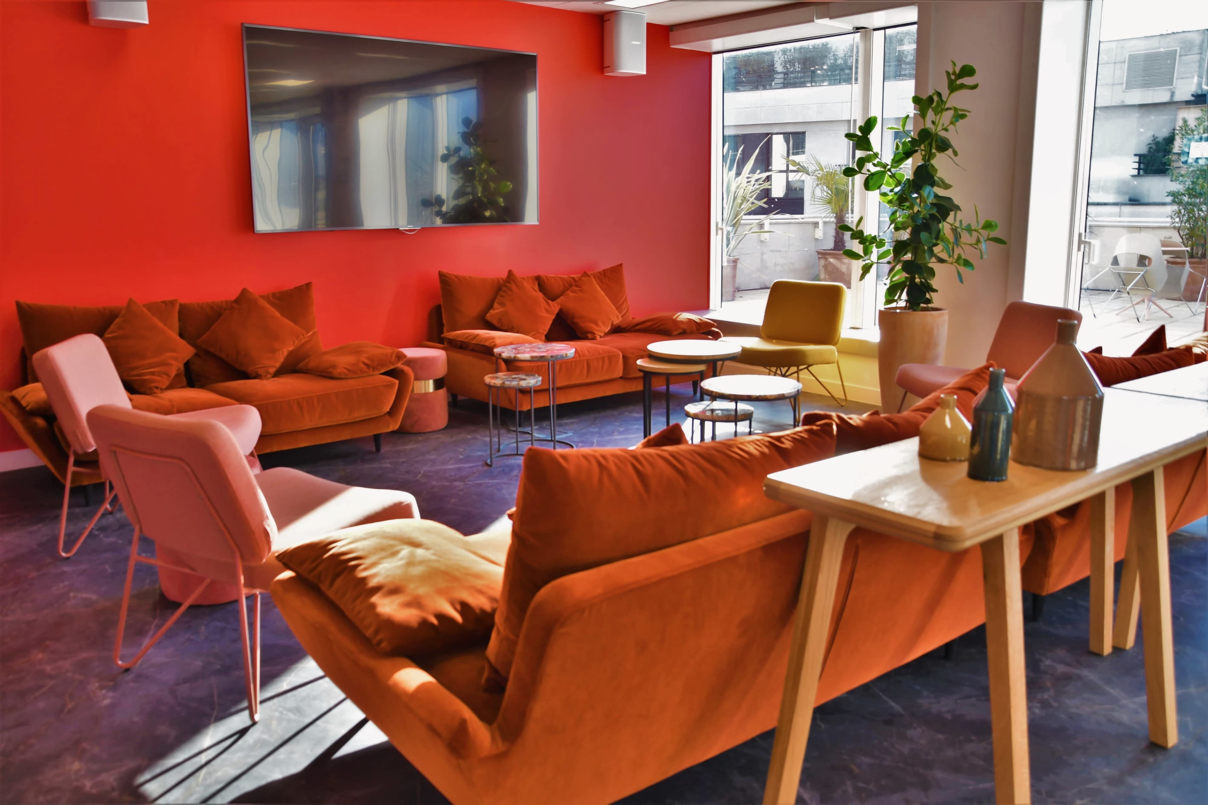 A modern lounge area with orange sofas and chairs arranged around small tables, set against a vibrant red wall and large windows.