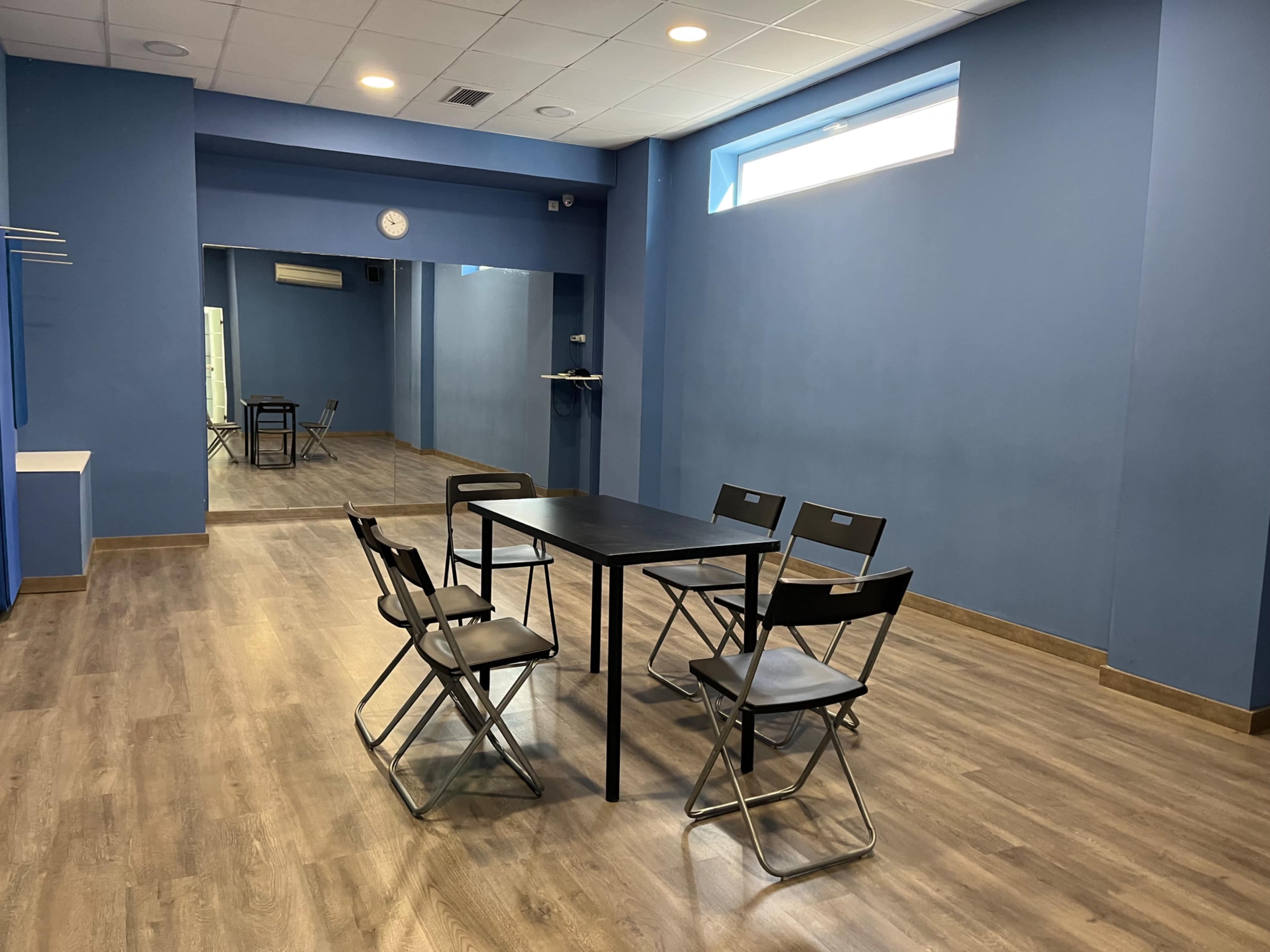 The image shows a room with blue walls featuring a table and six folding chairs arranged around it, with a mirror on the far wall.