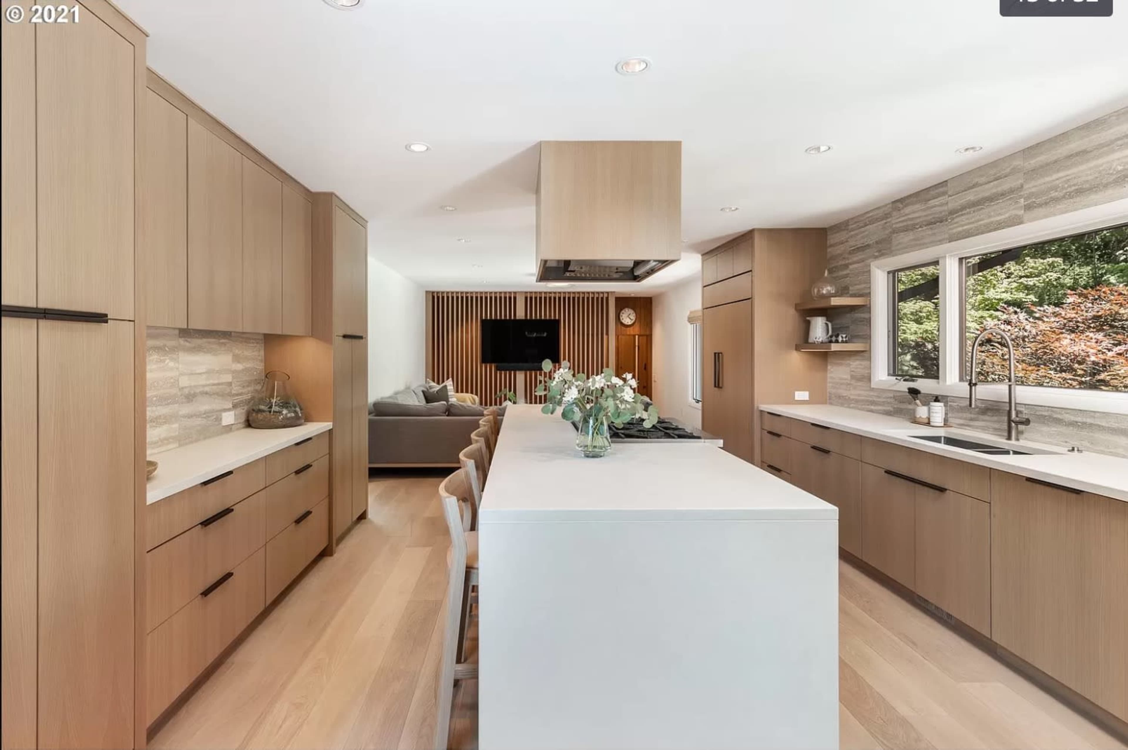 A modern kitchen features light wood cabinetry, a central island with seating, and a view of a living area with a gray sofa and stone accent wall.