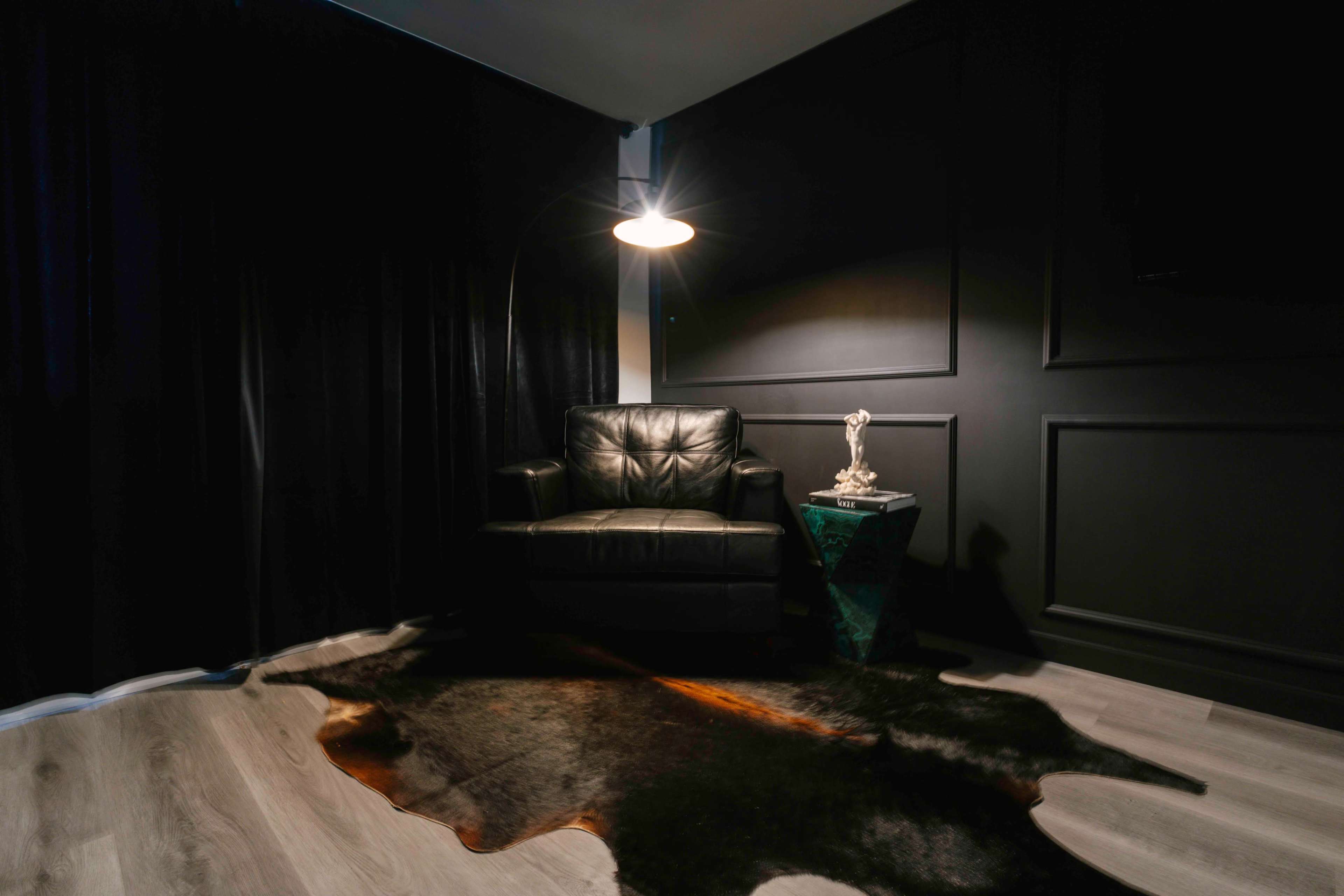 The image shows a dark room featuring a leather chair next to a small green side table, with a light fixture above and a textured rug on the floor.