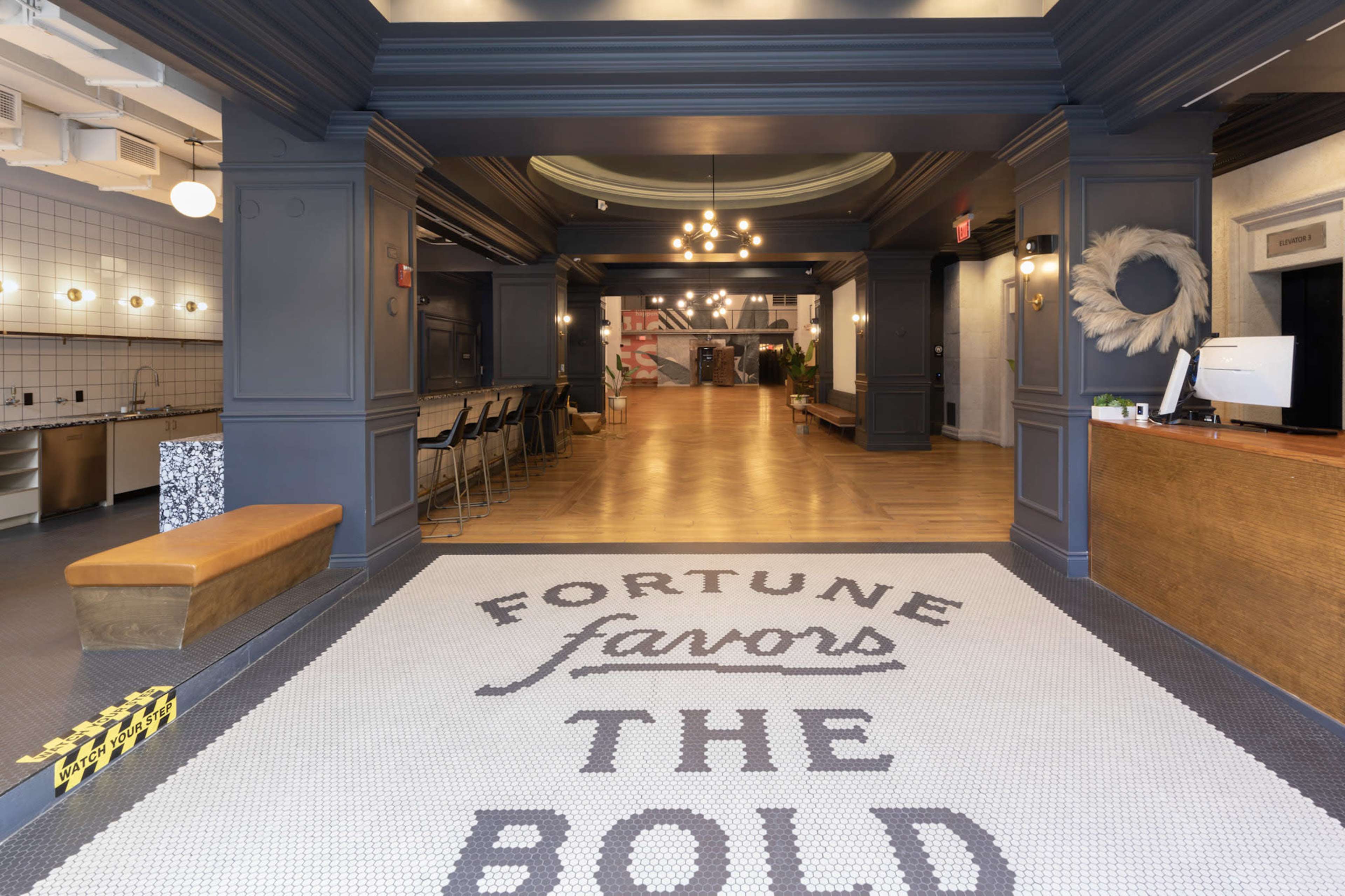 An indoor space featuring a large entrance mat with the words "FORTUNE favors THE BOLD," leading into a bright area with wooden flooring and modern decor.